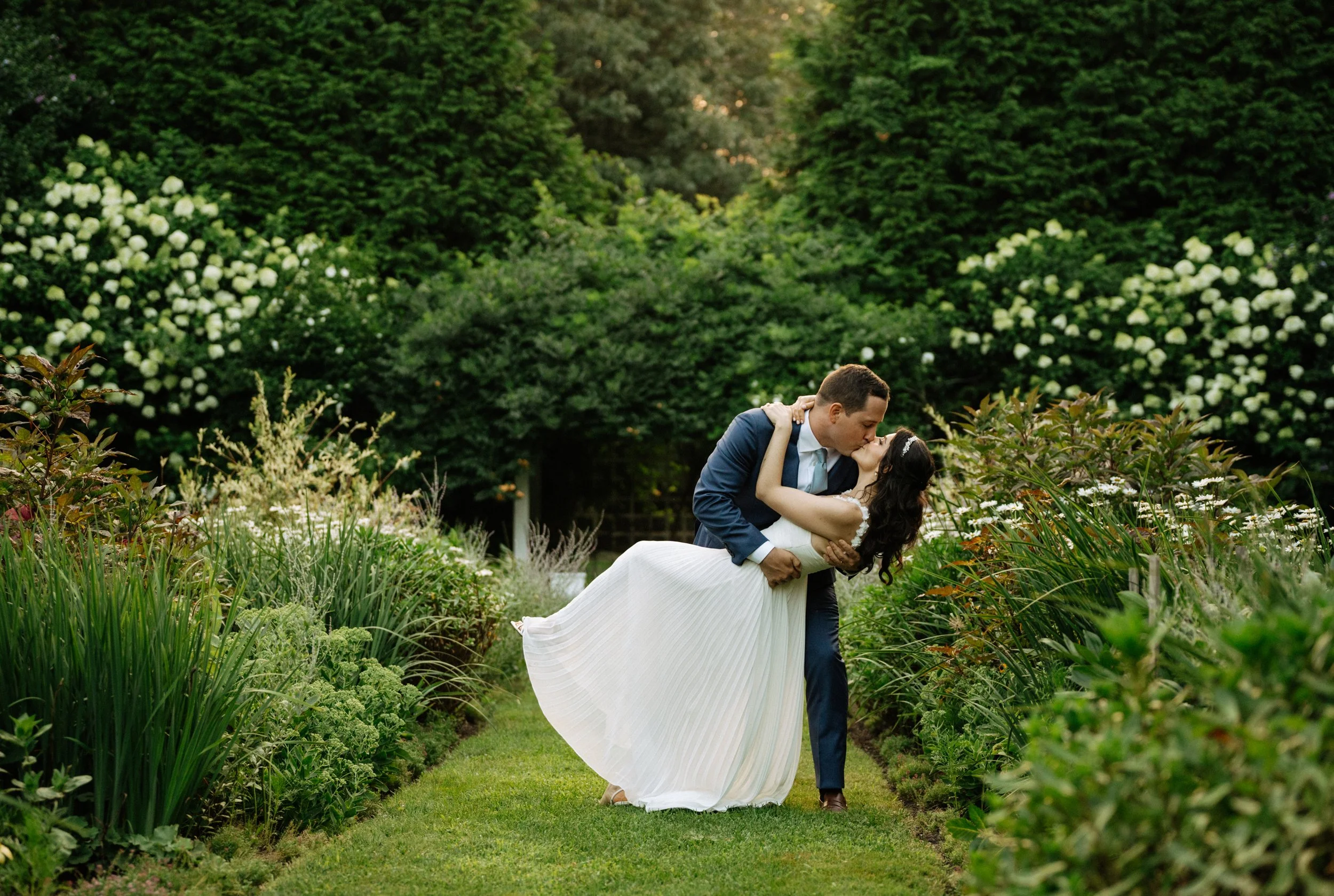 A groom dips and kisses a bride in a garden surrounded by lush greenery and blooming white flowers during a wedding.
