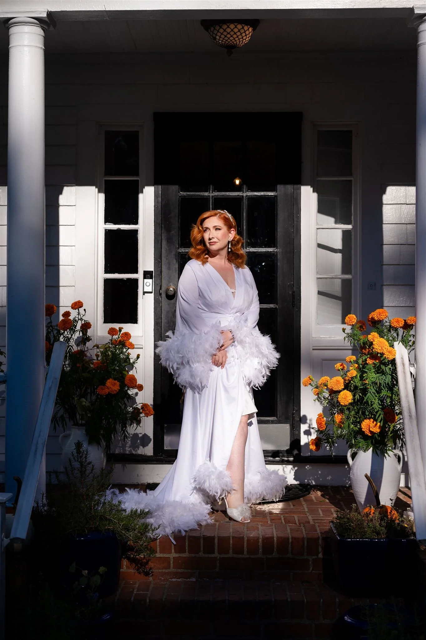 A woman with red hair standing on a porch in front of a black door at Lambert's Cove Inn on Martha's Vineyard, wearing a white dress with feathered embellishments, surrounded by orange flowers in pots, with columns and a small brick step.