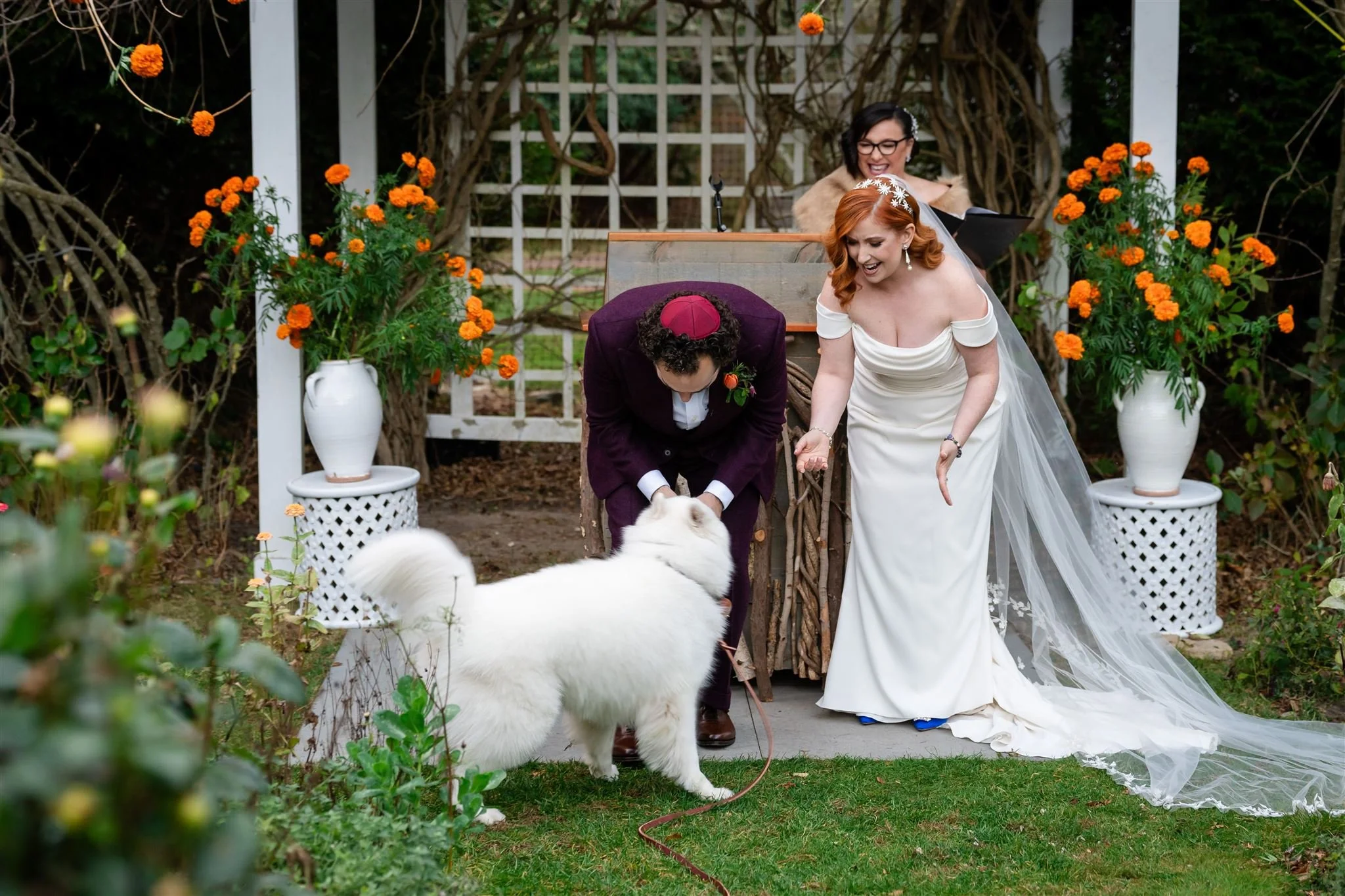 A bride and groom are getting married outdoors surrounded by orange flowers and greenery. The bride has red hair, is wearing a white off-shoulder wedding dress, and is smiling while holding the groom's hand. The groom is dressed in burgundy attire wi