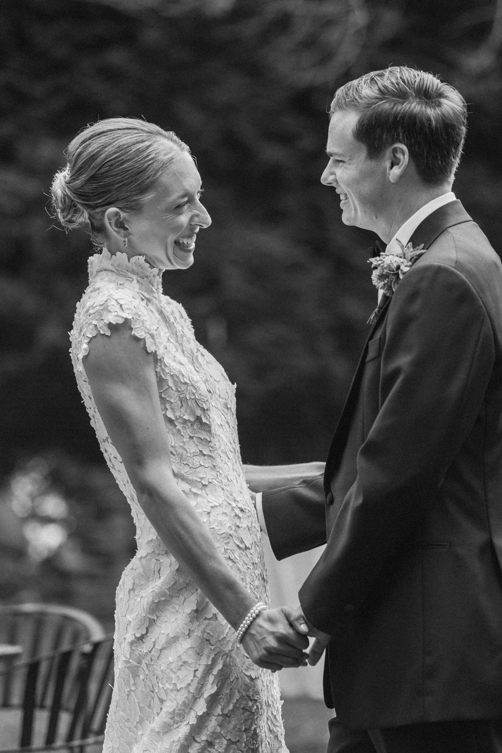 A black and white photo of a smiling bride and groom holding hands during their wedding ceremony at Lambert's Cove Inn. The bride wears a lace wedding dress and pearls, and the groom is in a tuxedo with a boutonnière. They face each other outdoors.