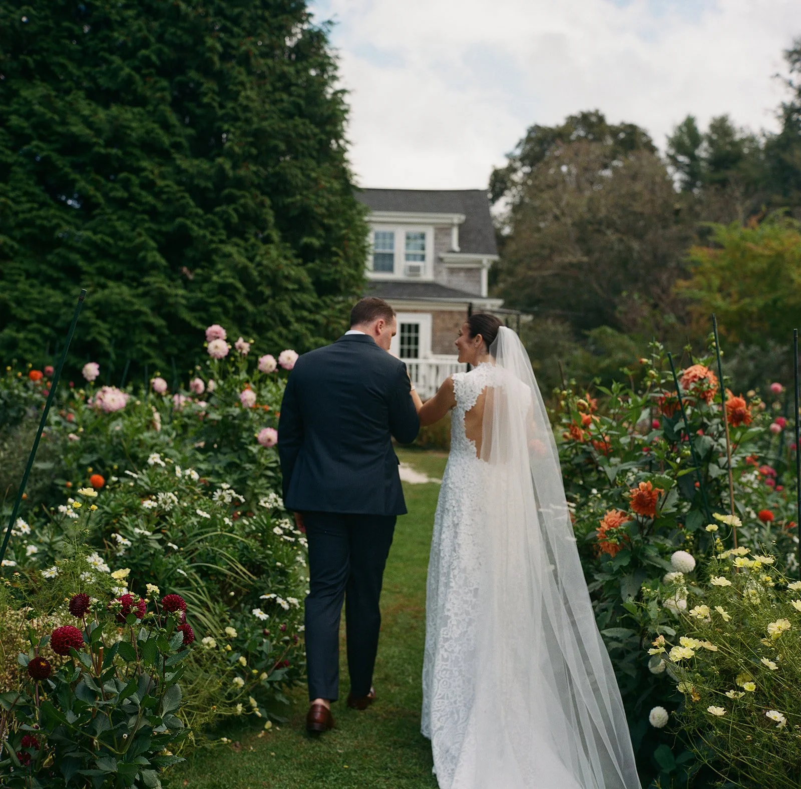 A bride and groom walking hand in hand through a garden filled with colorful flowers, with a house in the background, during a wedding reception at Lambert's Cove Inn on Martha's Vineyard.