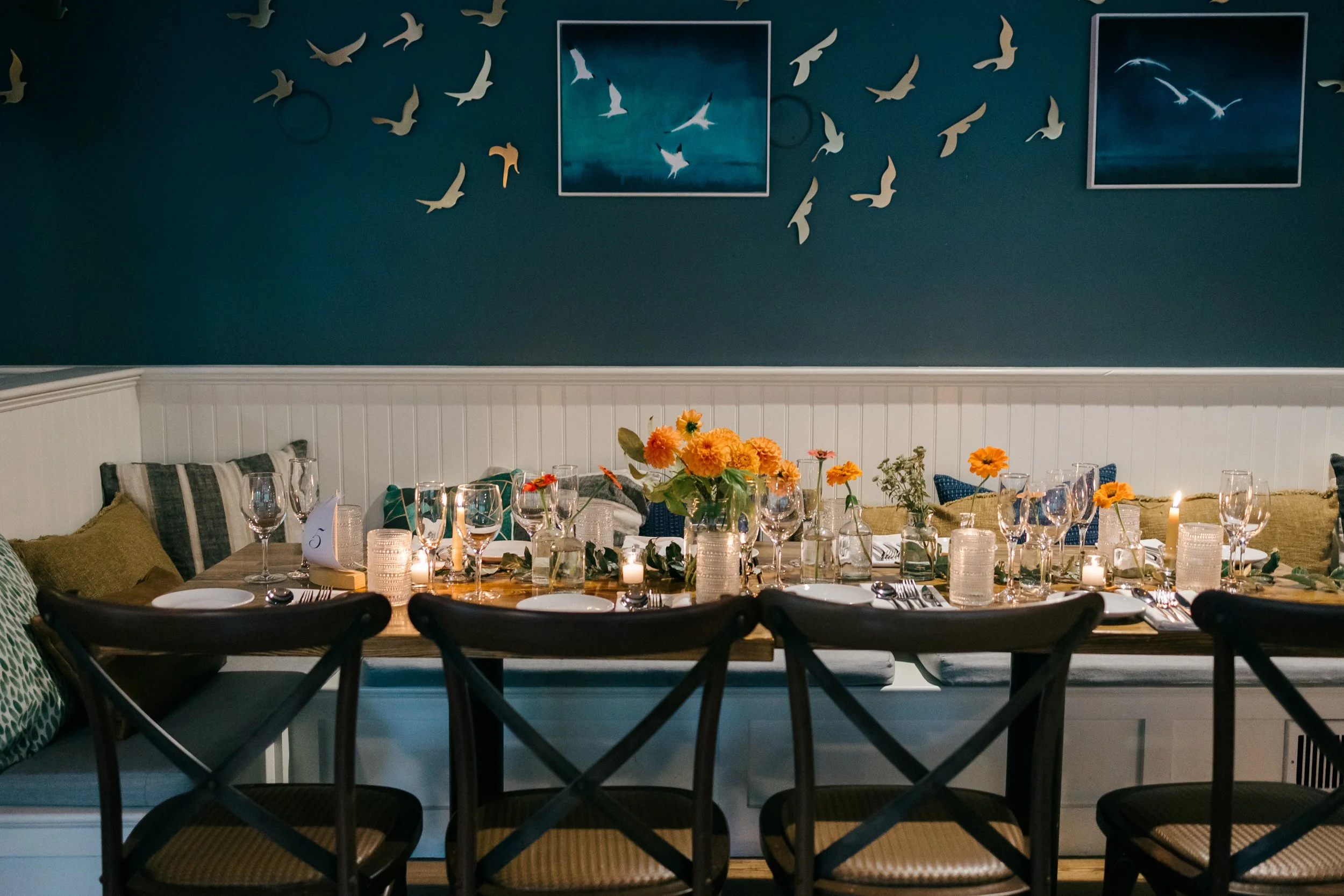 A dining room with a table set for a meal, decorated with flowers and candles, against a blue wall decorated with bird cutouts and framed pictures at Lambert's Cove Inn on Martha's Vineyard.