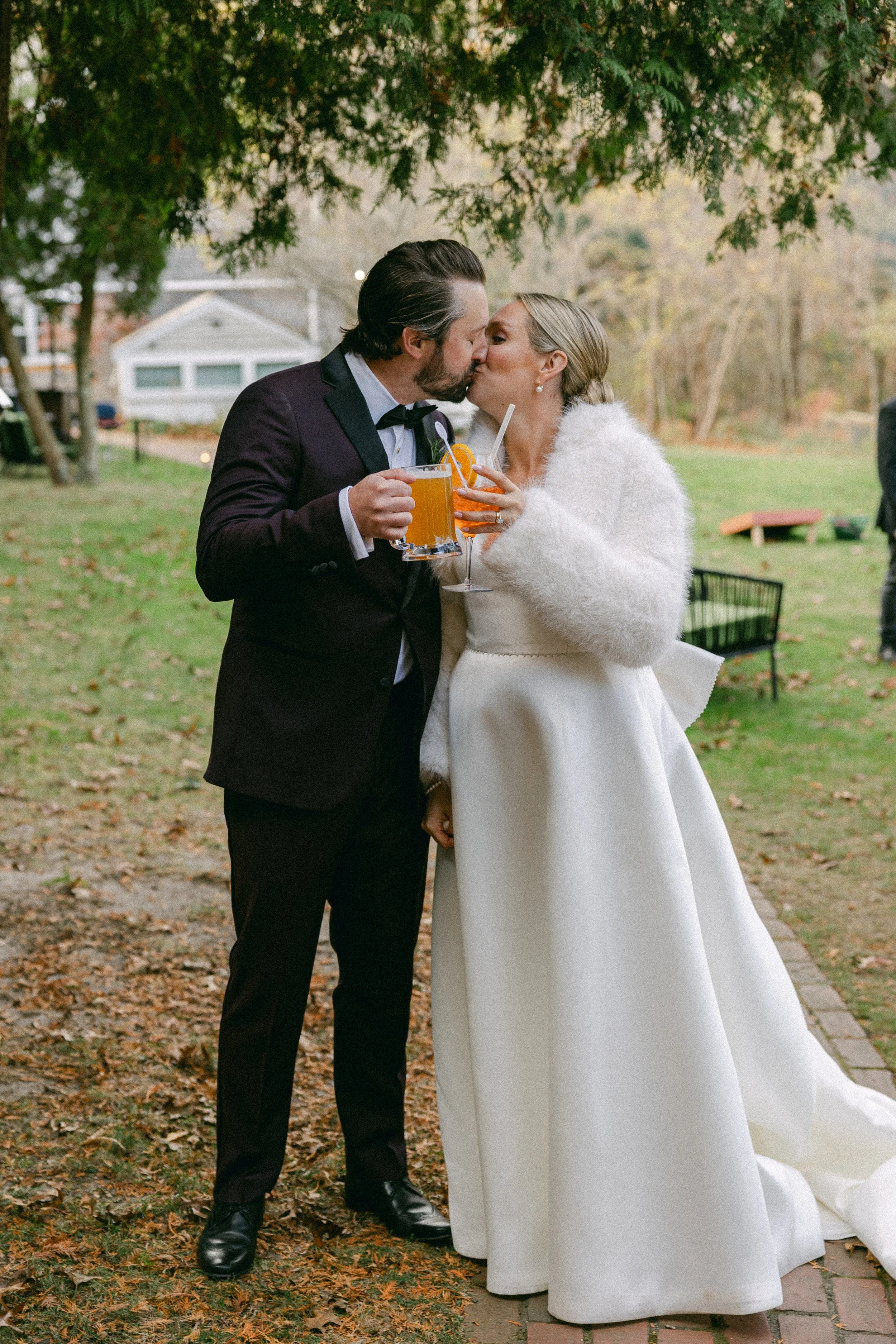 A couple dressed in wedding attire sharing a kiss outdoors. The man is in a tuxedo and the woman in a white wedding dress with a fluffy white jacket, holding drinks. The scene is set in a park with scattered leaves, trees, and a house in the backgrou