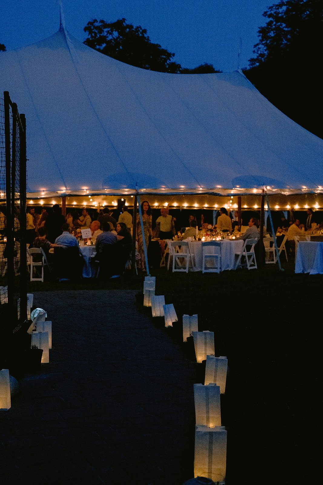 Outdoor evening event under a large canopy tent with string lights, people seated at round tables with white tablecloths, and a pathway lined with glowing paper lanterns.