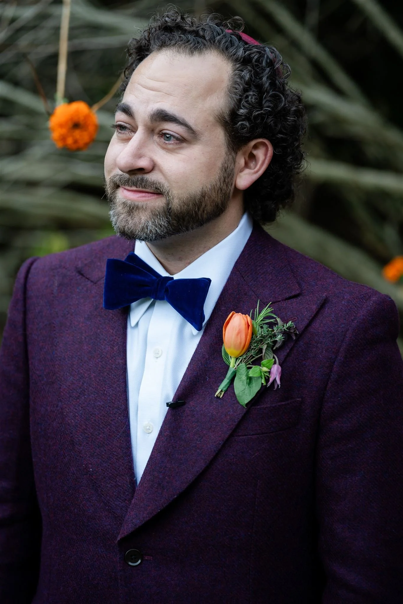 A man wearing a maroon blazer, white shirt, and a navy blue bow tie, with a boutonniere of orange and pink flowers on his left lapel, standing outdoors with greenery and orange flowers in the background.