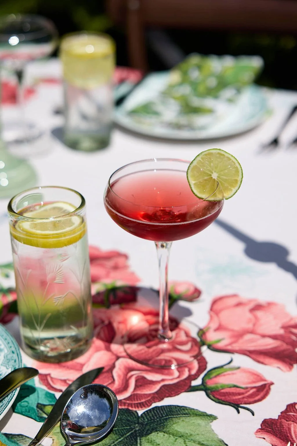 A table with pink and green floral tablecloth, set with cocktails garnished with lime slices, and dishes with utensils in the background.