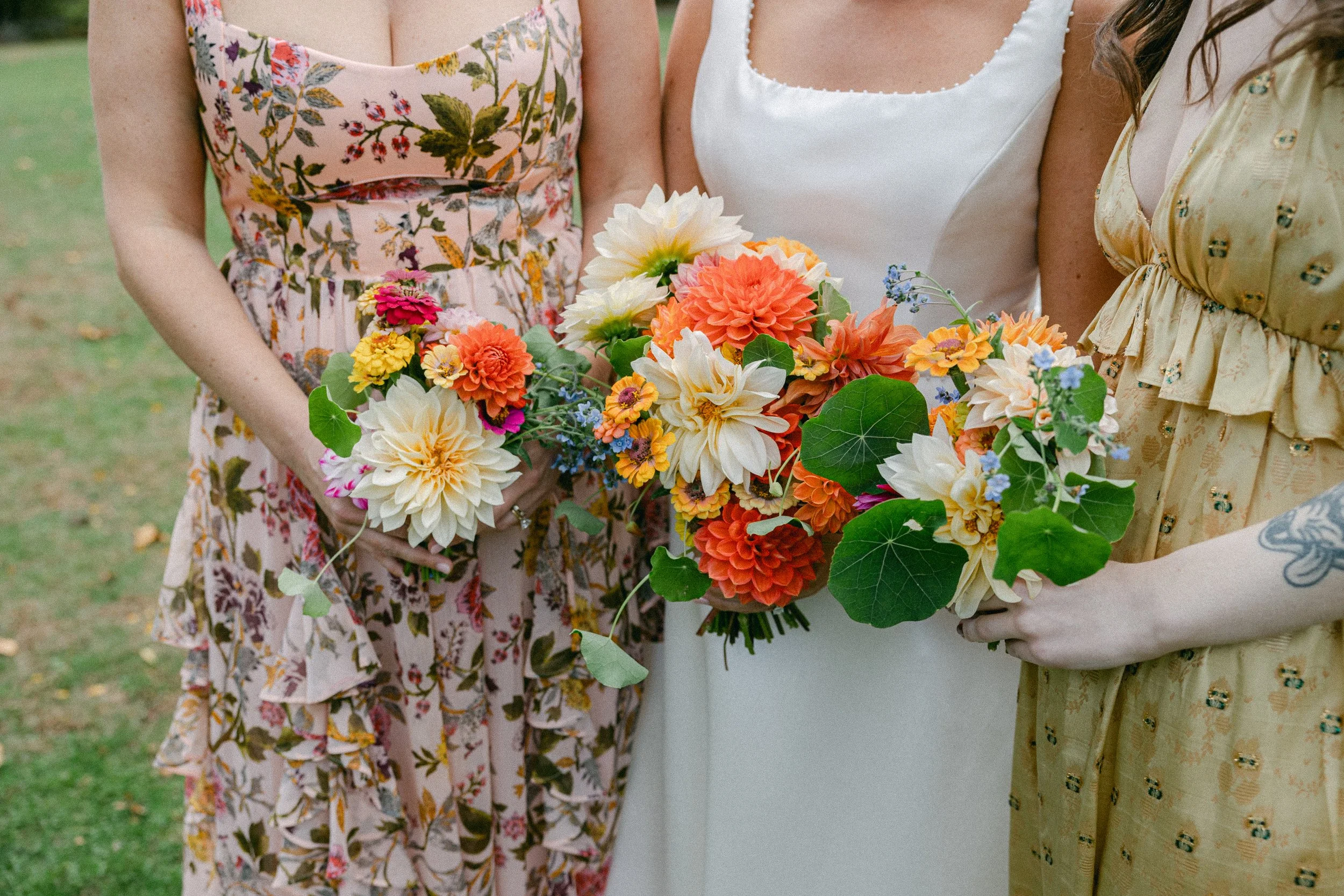Three women standing outdoors, holding a colorful bouquet of flowers, with parks and grass in the background during a wedding reception at Lambert's Cove Inn on Martha's Vineyard.