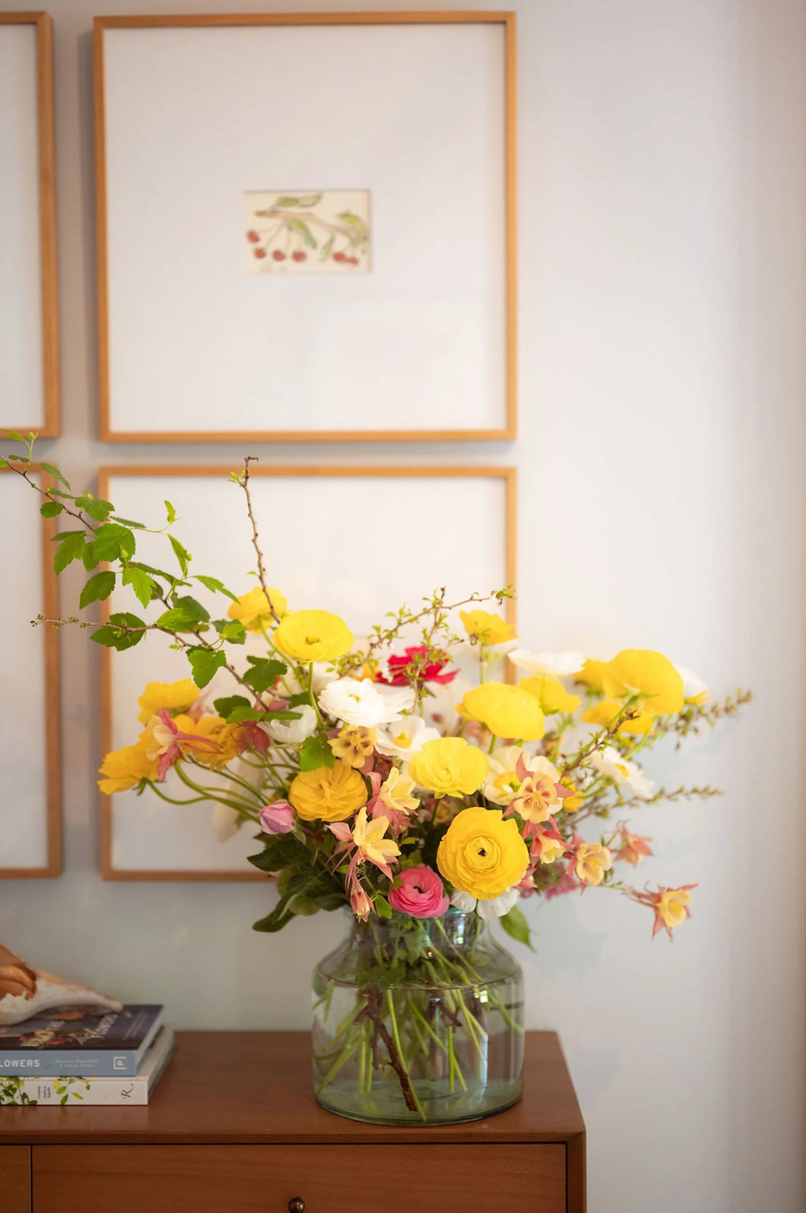 A large vase filled with a colorful assortment of fresh flowers sits on a wooden table. The background features a wall with four empty wooden picture frames and a small piece of artwork hanging. There are some books on the table next to the vase.