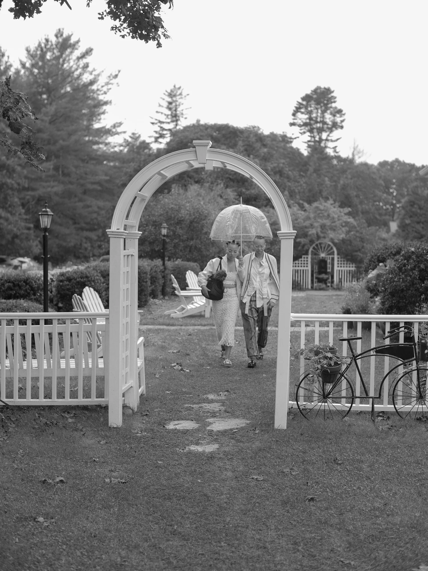 Two women holding an umbrella walking through a garden archway in black and white during a wedding at Lambert's Cove Inn. The garden is landscaped with trees, shrubs, and benches, and a bicycle with a flower pot is parked on the right.