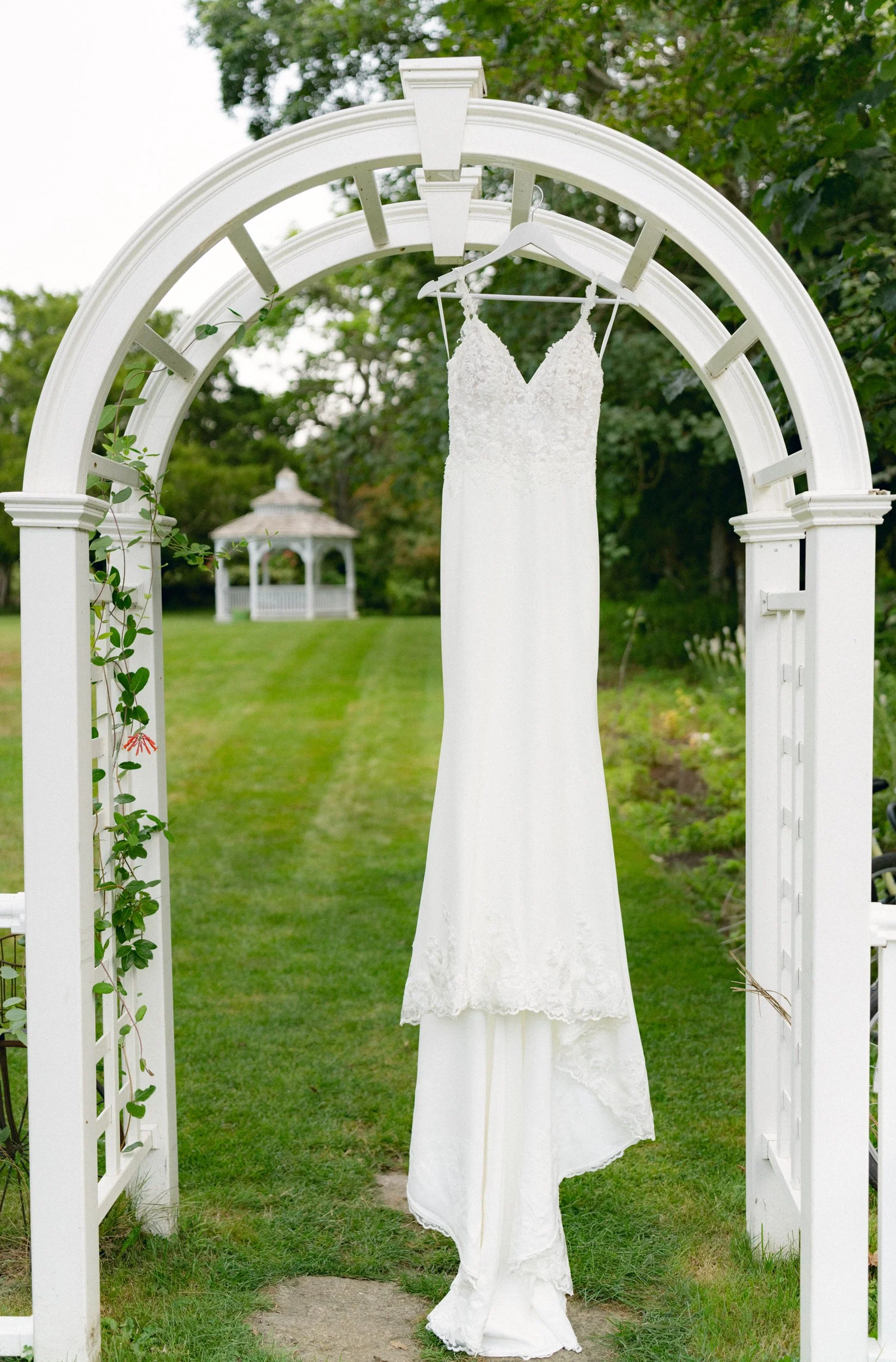 White wedding dress hanging on a hanger on a white archway in a garden setting with a green lawn and trees in the background at Lambert's Cove Inn on Martha's Vineyard.
