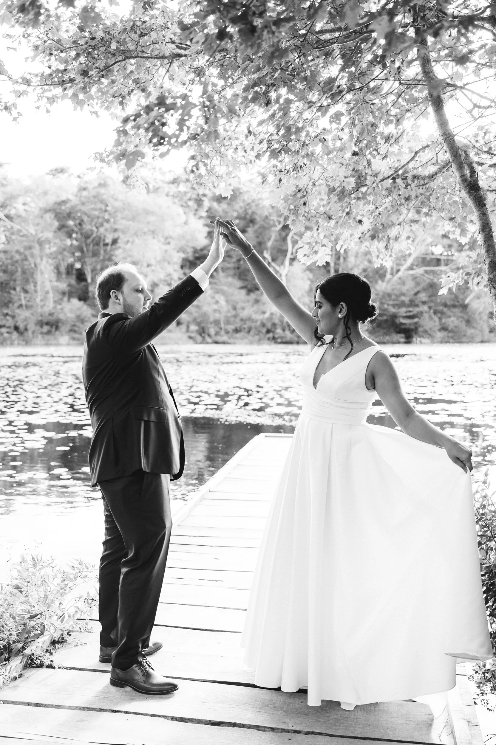 A black and white photo of a bride and groom standing on a dock by a river, touching hands under a tree with leaves, surrounded by nature at Lambert's Cove Inn on Martha's Vineyard.