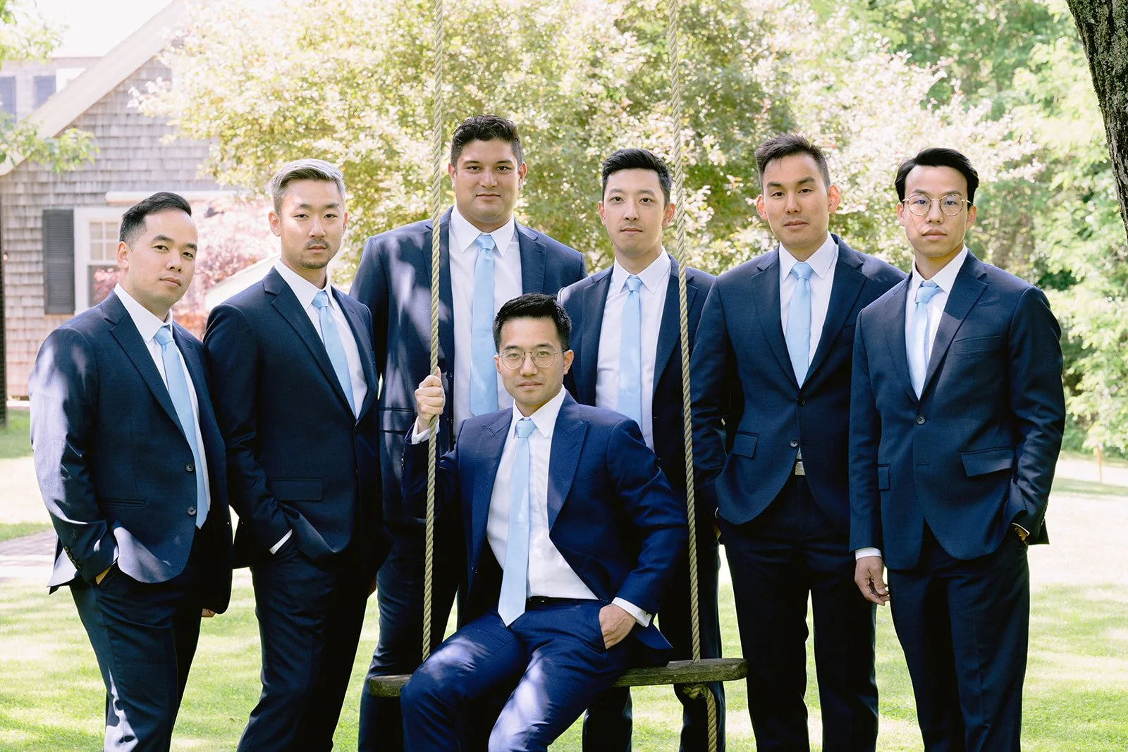 Group of eight men dressed in navy wedding suits and light blue ties, standing outdoors on grass with trees during a wedding at Lambert's Cove Inn on Martha's Vineyard. One man is sitting on a swing, and another is holding the swing's rope.