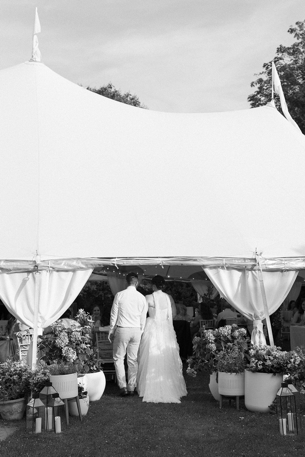 Black and white photo of a bride and groom walking into a wedding reception at Lambert's Cove Inn on Martha's Vineyard under a large white canopy decorated with flowers and lights, with guests seated at tables in the background.