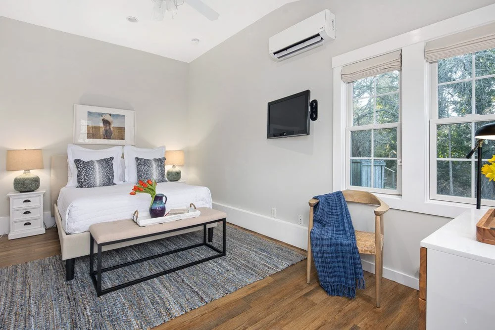 A bedroom with a bed, two nightstands, lamps, a painting, a bench, a chair with a blue blanket, a dresser, a window, and an air conditioner.
