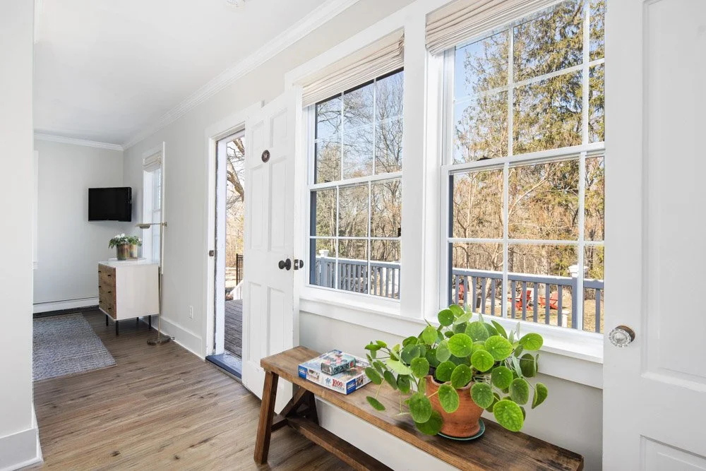 Bright living room with large windows, a small wooden bench with a potted plant and board games, and a deck outside.