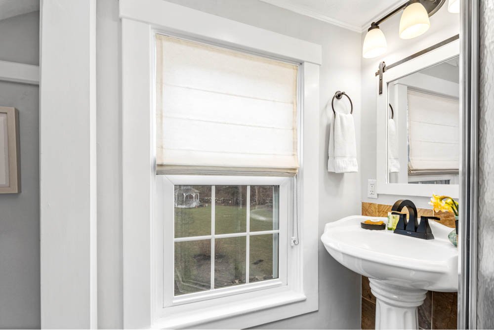 A small bathroom with a white pedestal sink, a mirror with lighting, a towel ring with a white towel, a large window with a blind, and some small decorative items on the sink.