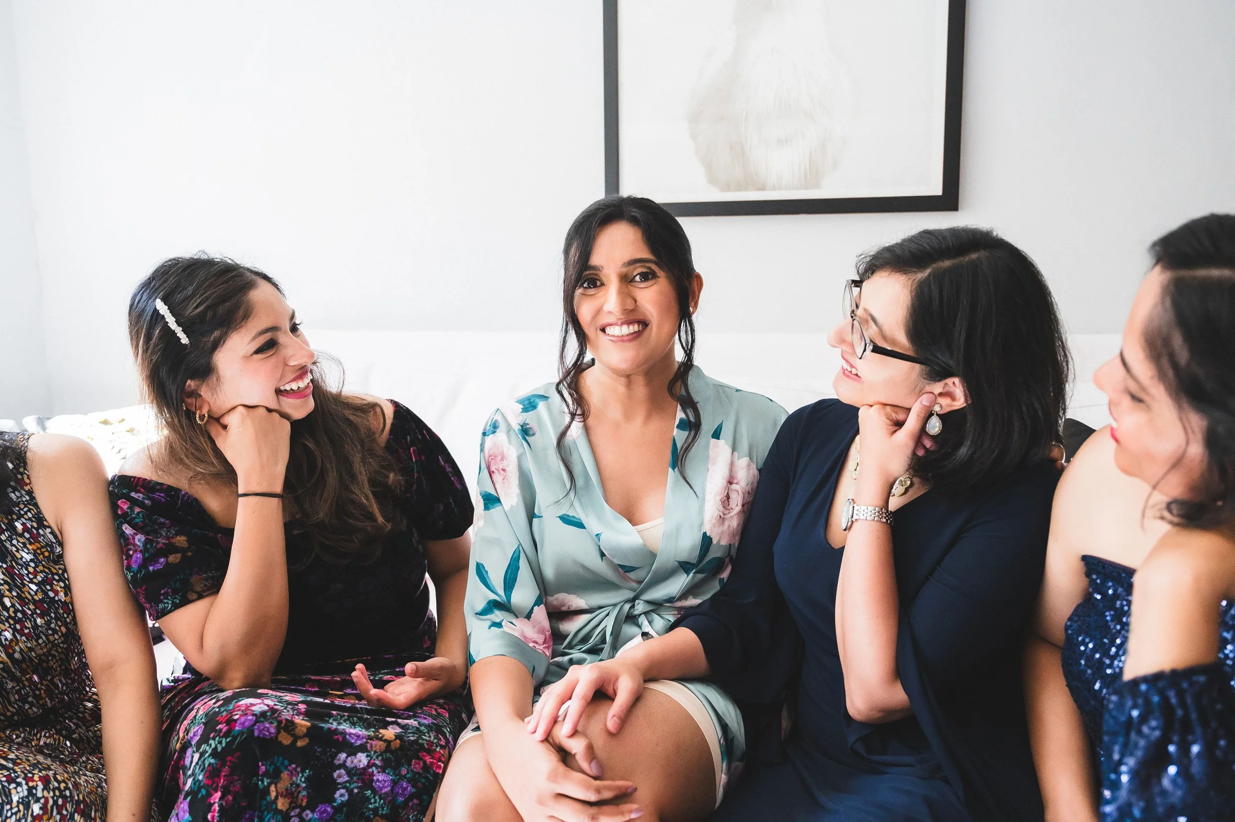 Group of women sitting together, smiling, and engaged in conversation in a bright, modern room with white walls and a framed artwork.