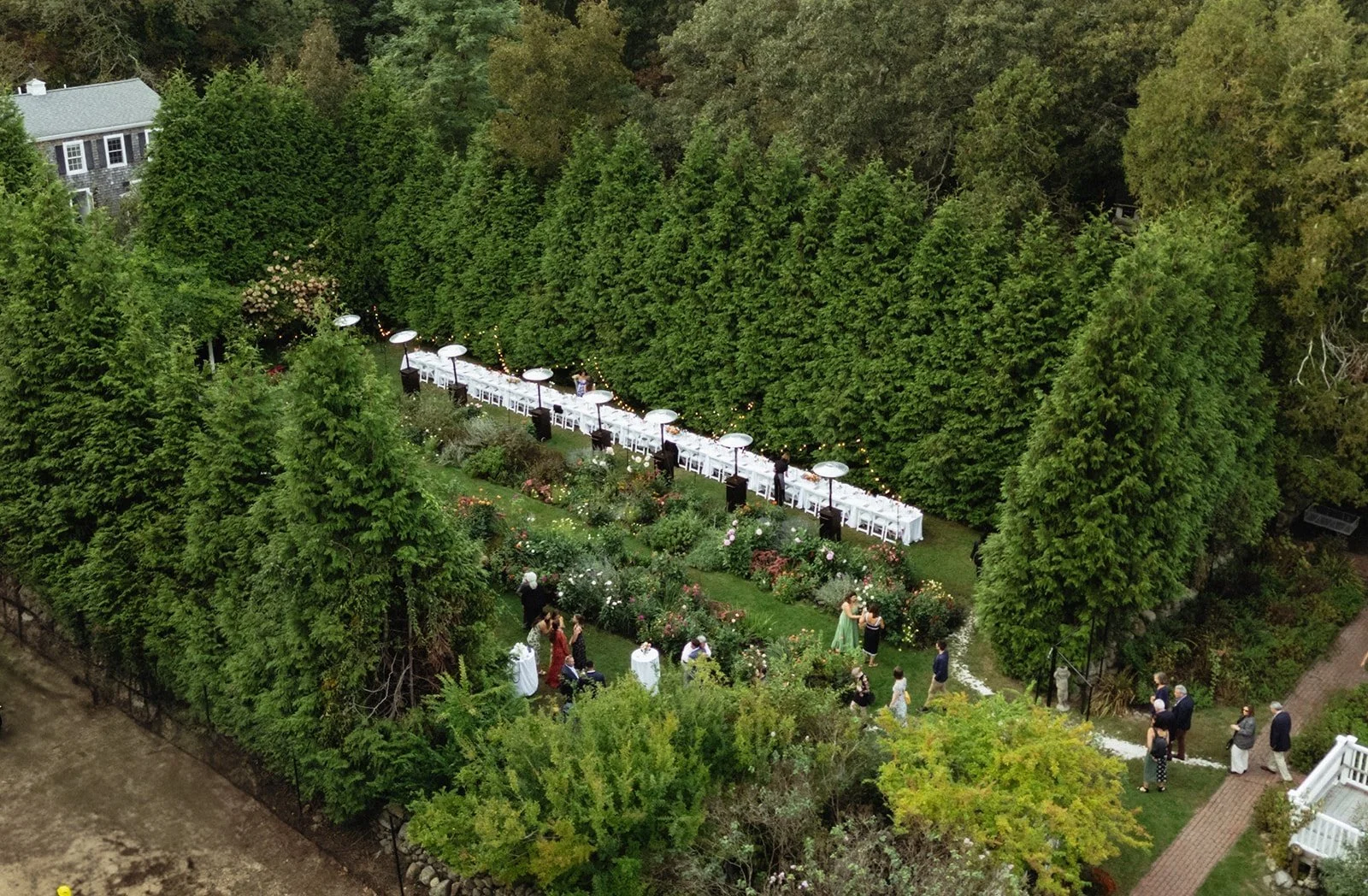 An outdoor garden wedding reception area with a long table under umbrellas, surrounded by lush green trees and flowers, with guests mingling.