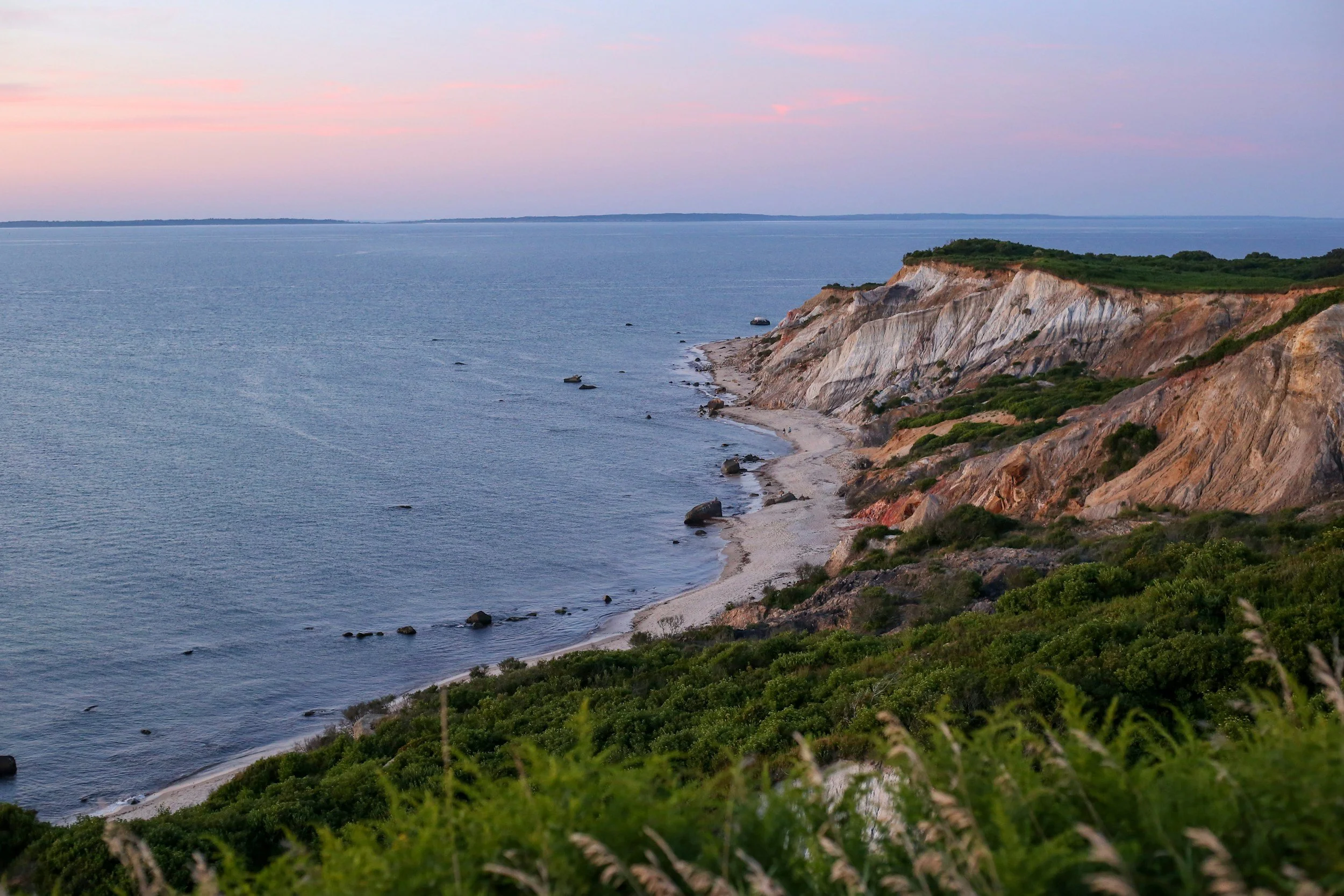 Cliffside overlooking a calm ocean with pink and blue sky at sunset, green vegetation on the cliff, and rocky shoreline below on Martha's Vineyard.