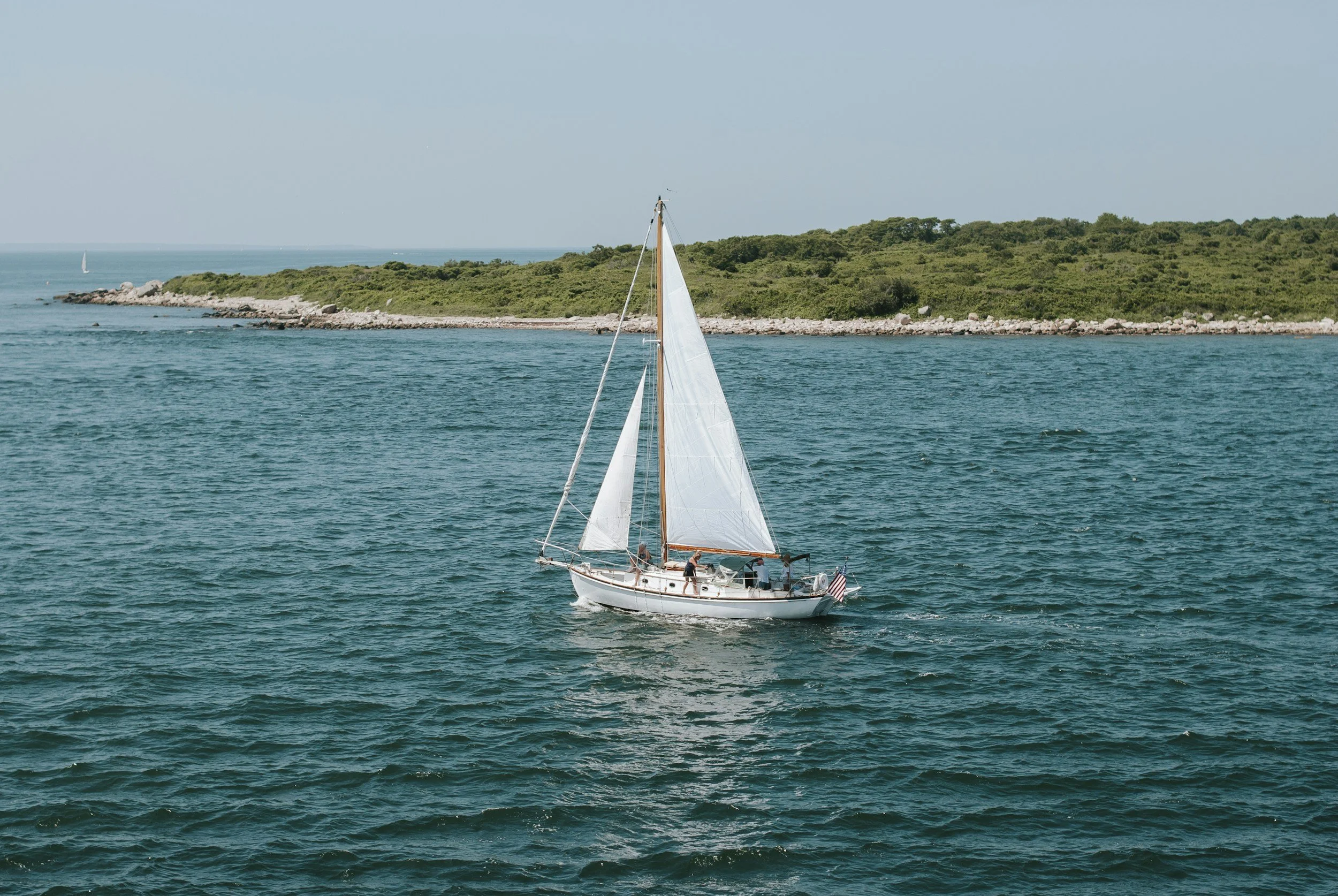 A sailboat with white sails on the water near the shoreline of Martha's Vineyard under a clear blue sky.