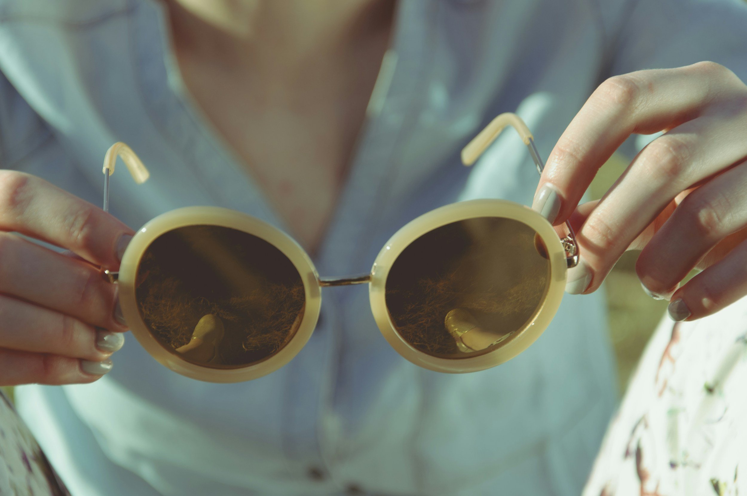 A person with light-colored nails holding a pair of yellow sunglasses with circular dark lenses, sunglasses positioned close to the camera lens