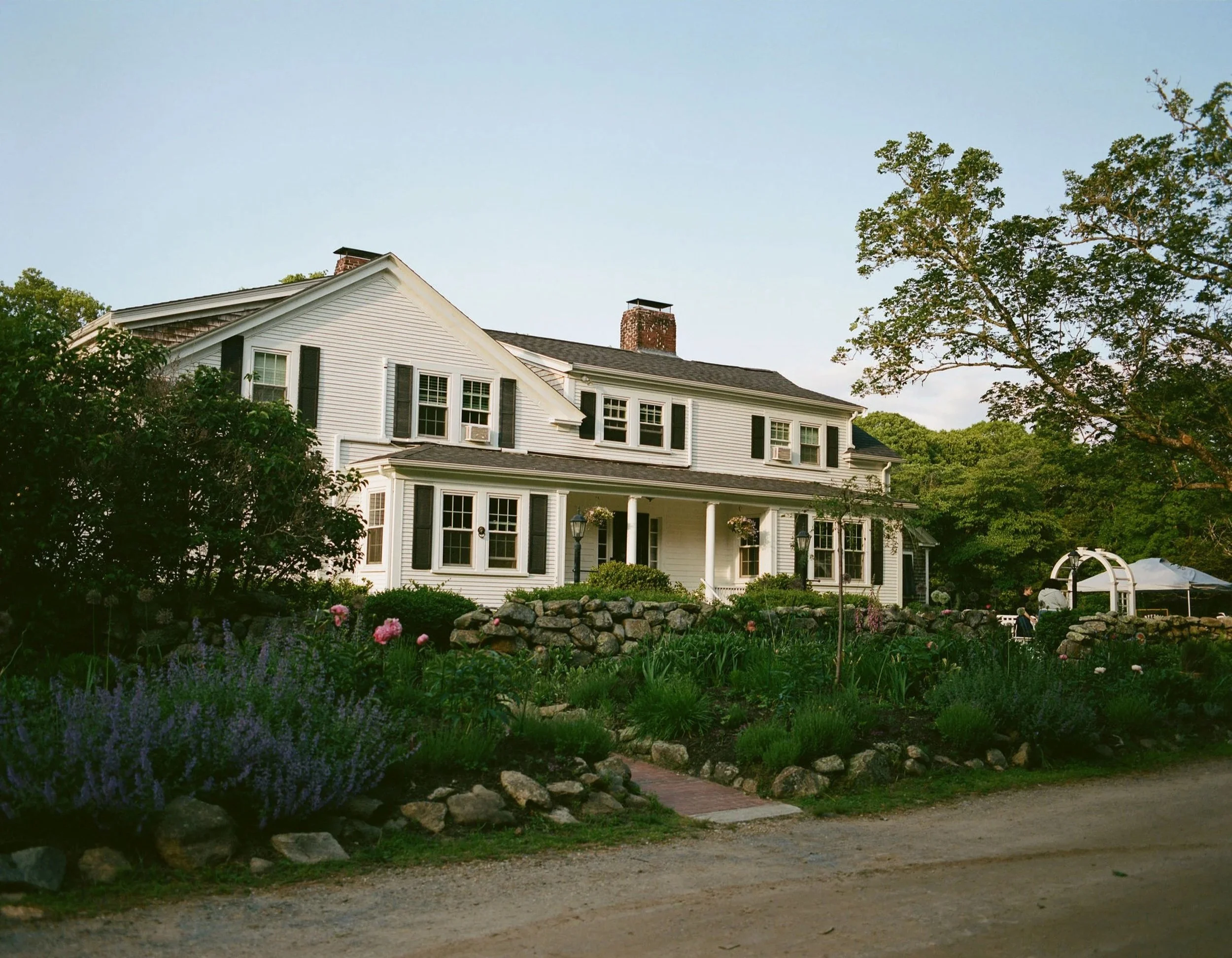 A large white house with black shutters and multiple windows, surrounded by trees and a garden in the foreground, with a gravel path in front and a blue sky above.