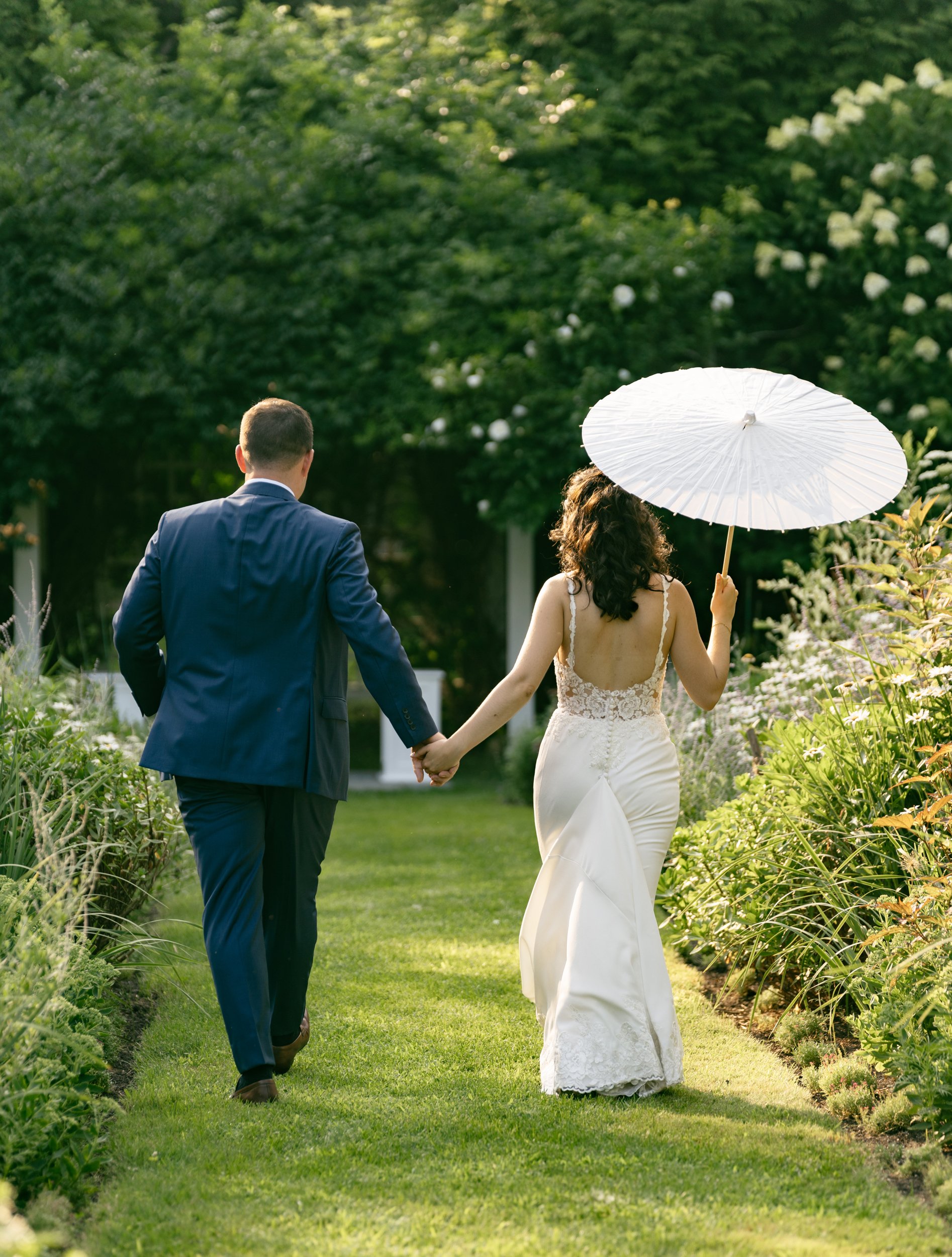 A couple walking hand in hand in the English garden at Lambert's Cove Inn on Martha's Vineyard during their wedding, the woman holding a white parasol. The woman wears a lacy white wedding dress, and the man wears a dark blue suit.