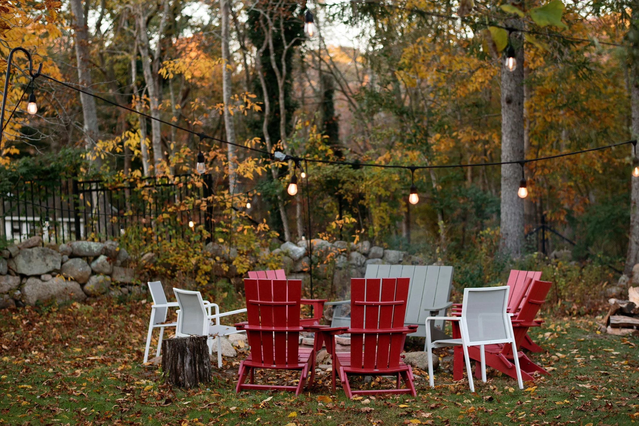 Colorful outdoor seating area at Lambert's Cove Inn with red and white chairs, surrounded by trees with autumn leaves, string lights hanging overhead, stone wall in background, and a rocky ground with fallen leaves.