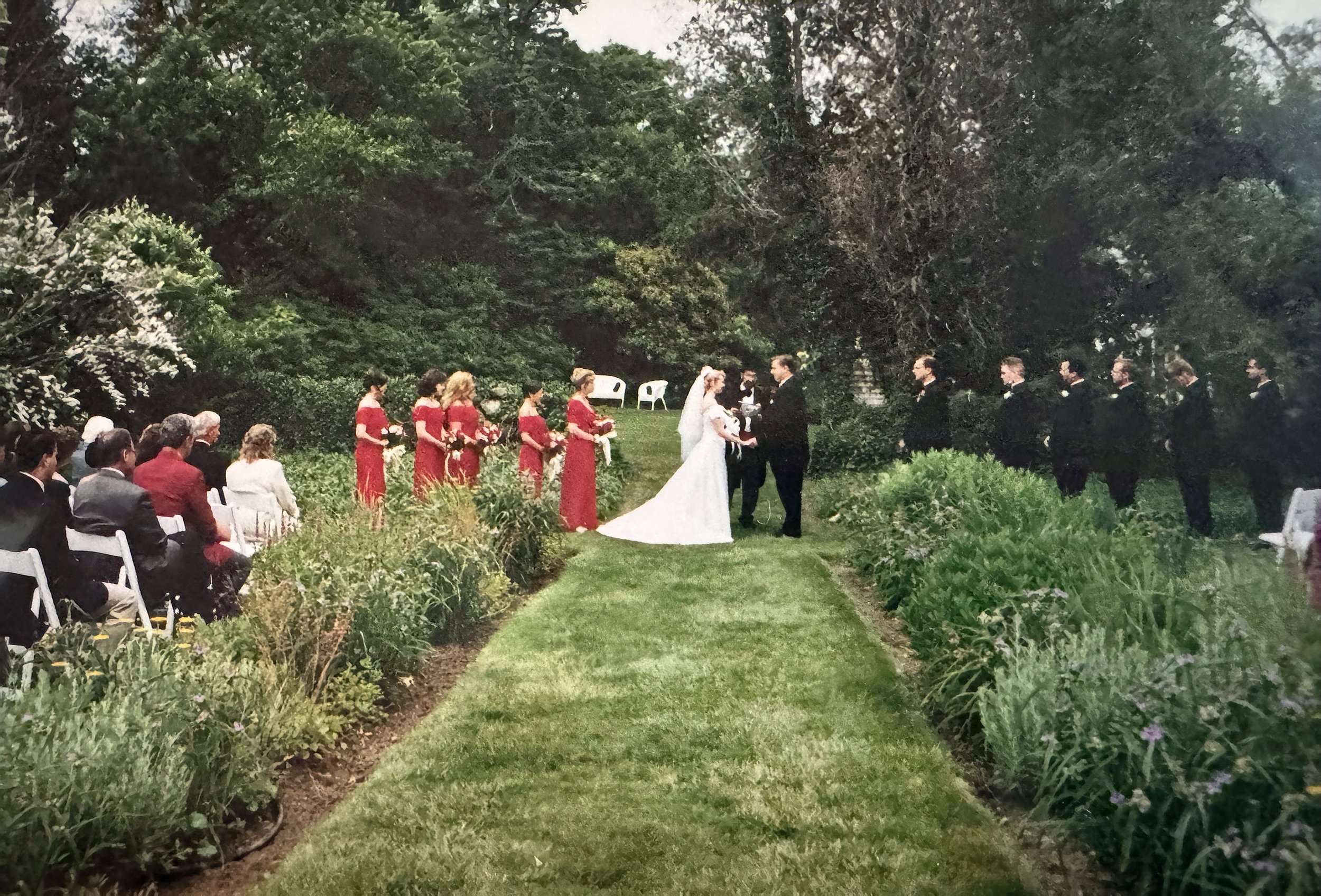 A wedding ceremony taking place outdoors surrounded by trees and greenery. The bride and groom are exchanging vows, with bridesmaids in red dresses on one side and groomsmen in black suits on the other.