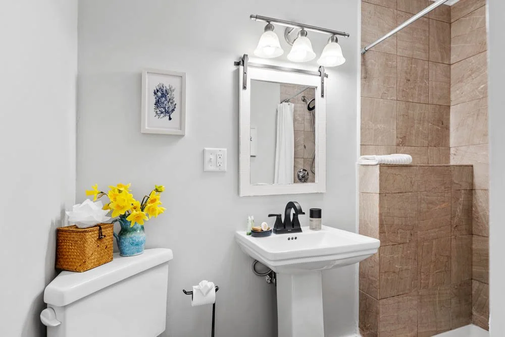 Bathroom with white walls, a mirror above a pedestal sink, a shower with brown tiles, a small framed floral art, a vase of yellow flowers, tissue box on toilet tank, and a towel on shower wall.