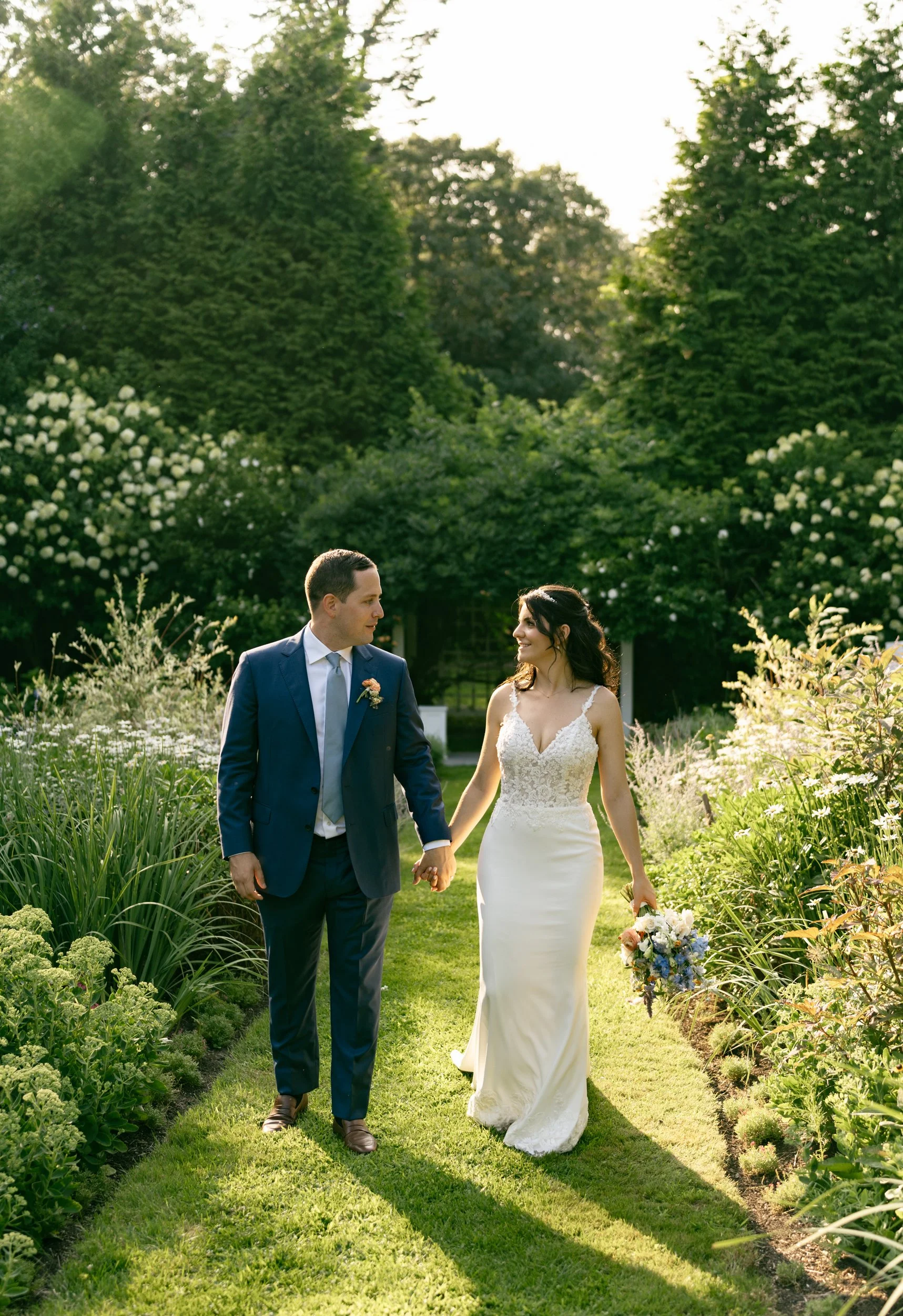 A newly married couple walking hand in hand through the English garden at Lambert's Cove Inn on Martha's Vineyard with lush greenery and blooming plants.