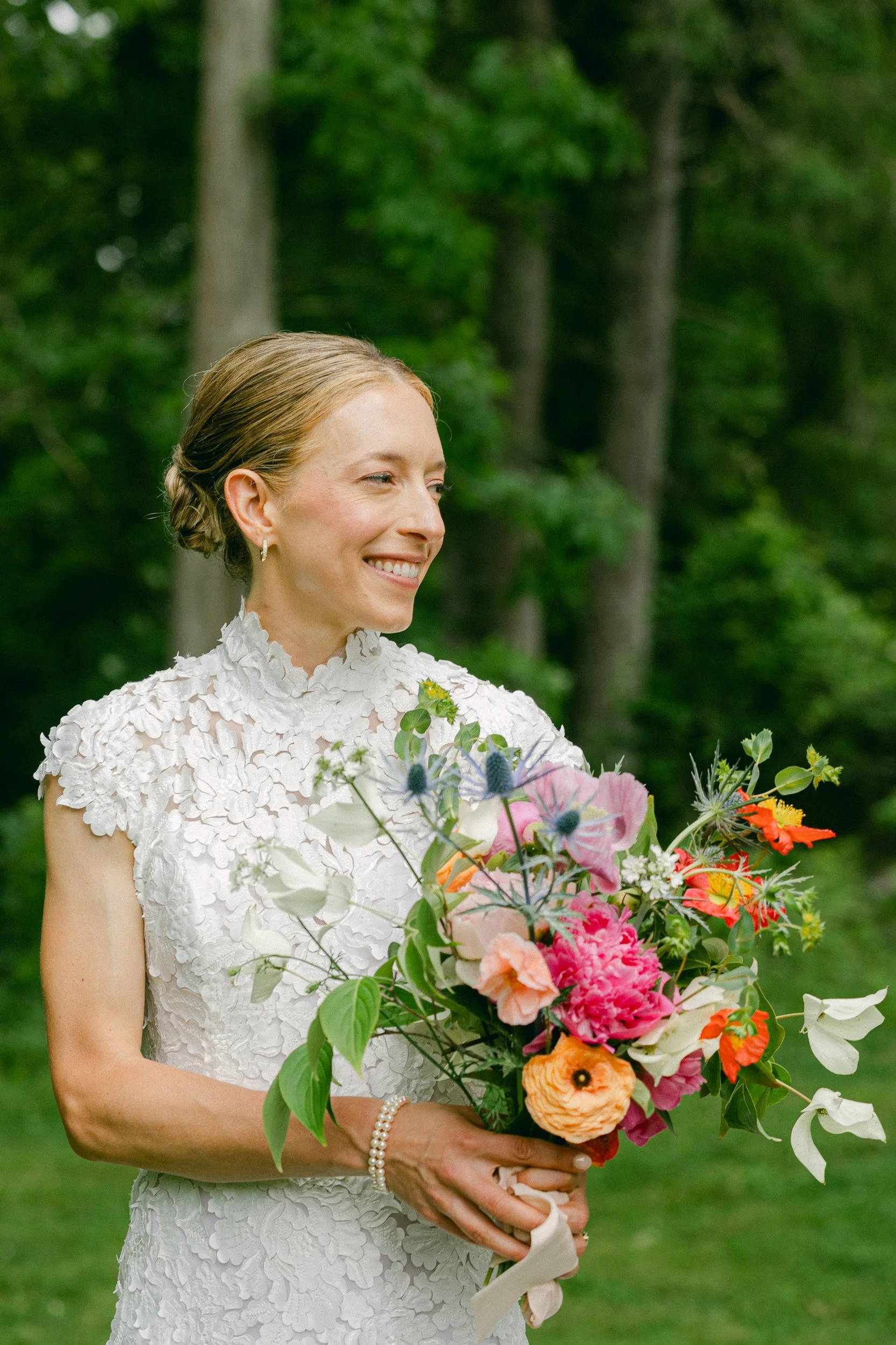 A bride in a white dress holding a colorful bouquet outdoors, with green trees in the background, during a wedding at Lambert's Cove Inn on Martha's Vineyard.