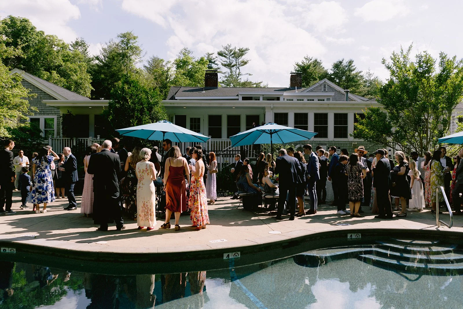 People gathered for an outdoor wedding reception at Lambert's Cove Inn on Martha's Vineyard at a backyard patio with umbrellas and pool, house in background, in sunny weather.