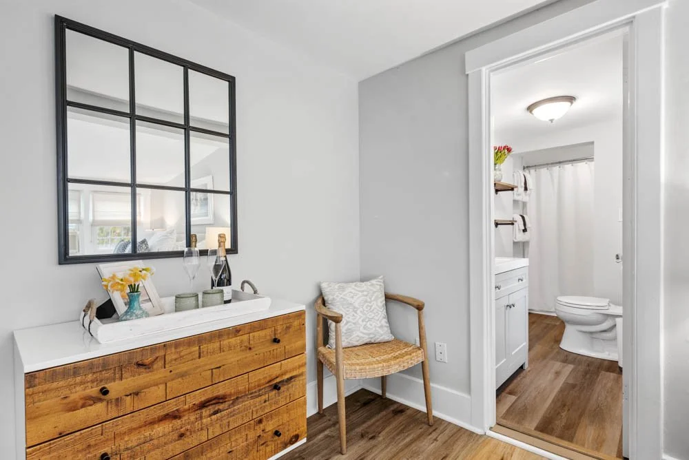 Interior of a light and airy home bathroom area with a wooden dresser, a mirror, a chair, and a view of the bathroom with a vanity and toilet.
