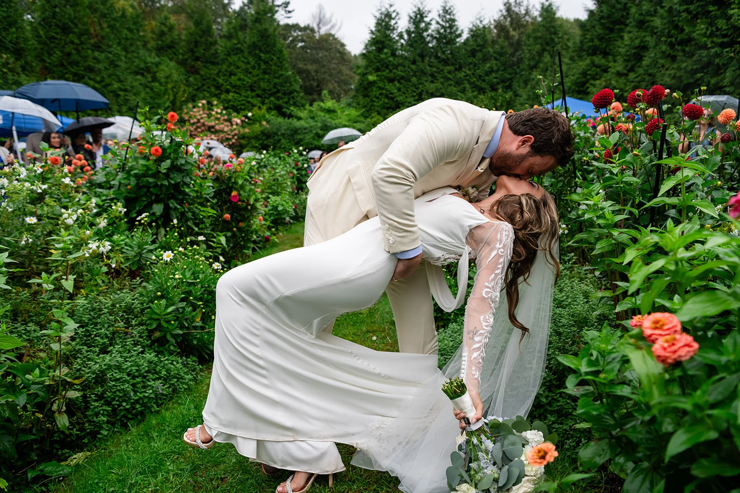 A newlywed couple sharing a kiss outdoors during a wedding ceremony in a garden, surrounded by flowers and onlookers holding umbrellas.