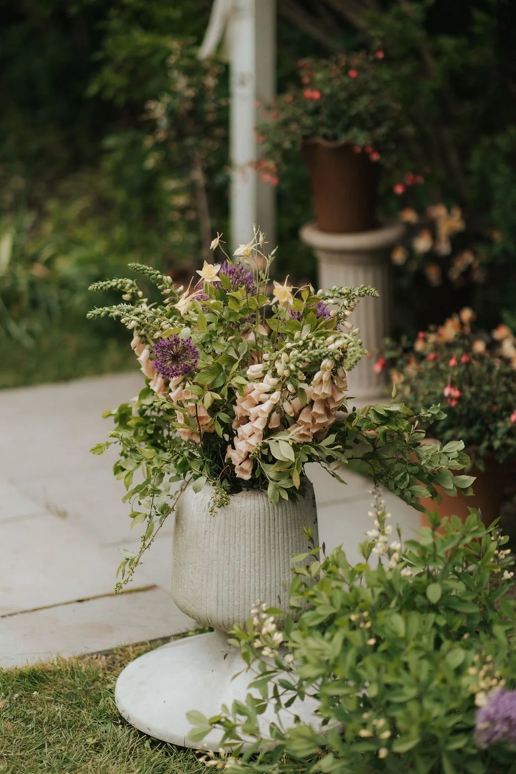A flower arrangement in a round, ribbed ceramic vase on a white stand outdoors, with a background of greenery and other potted plants.