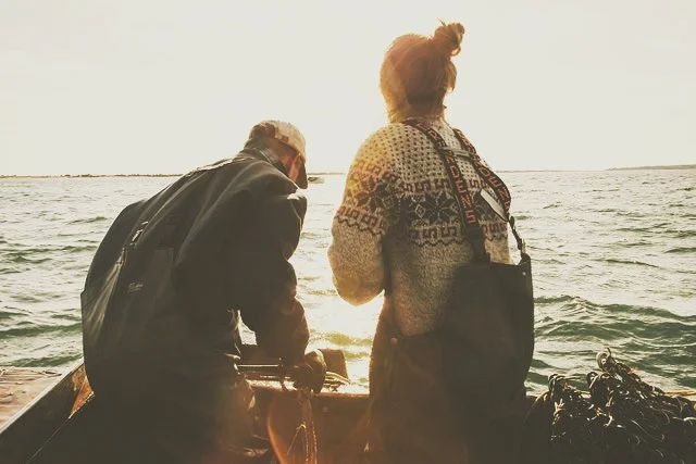 Two people on a boat fishing in the ocean at sunset, one older man and one woman with a bun hairstyle, both wearing jackets.