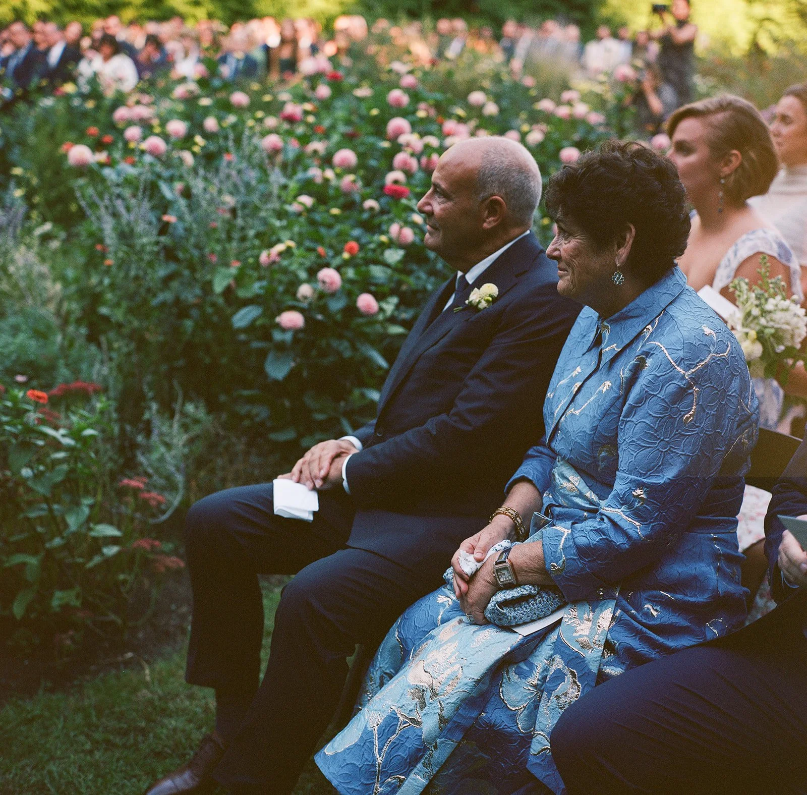 A group of people attending an outdoor event sitting on benches among flowering plants, with a focus on an elderly woman in a blue patterned dress and a man in a dark suit sitting beside her uring a wedding at Lambert's Cove Inn on Martha's Vineyard.