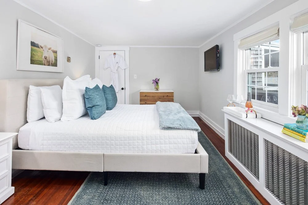 Bedroom with white bed, blue pillows, wall art of a sheep, wooden dresser with flowers, wall-mounted TV, window with blinds, and wine bottle with glasses on windowsill.