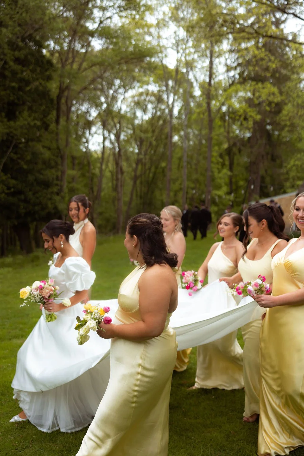Bride and bridesmaids in yellow dresses walking outdoors at Lambert's Cove Inn on Martha's Vineyard through a green wooded area with trees, carrying bouquets of pink, yellow, and white flowers for a wedding procession.