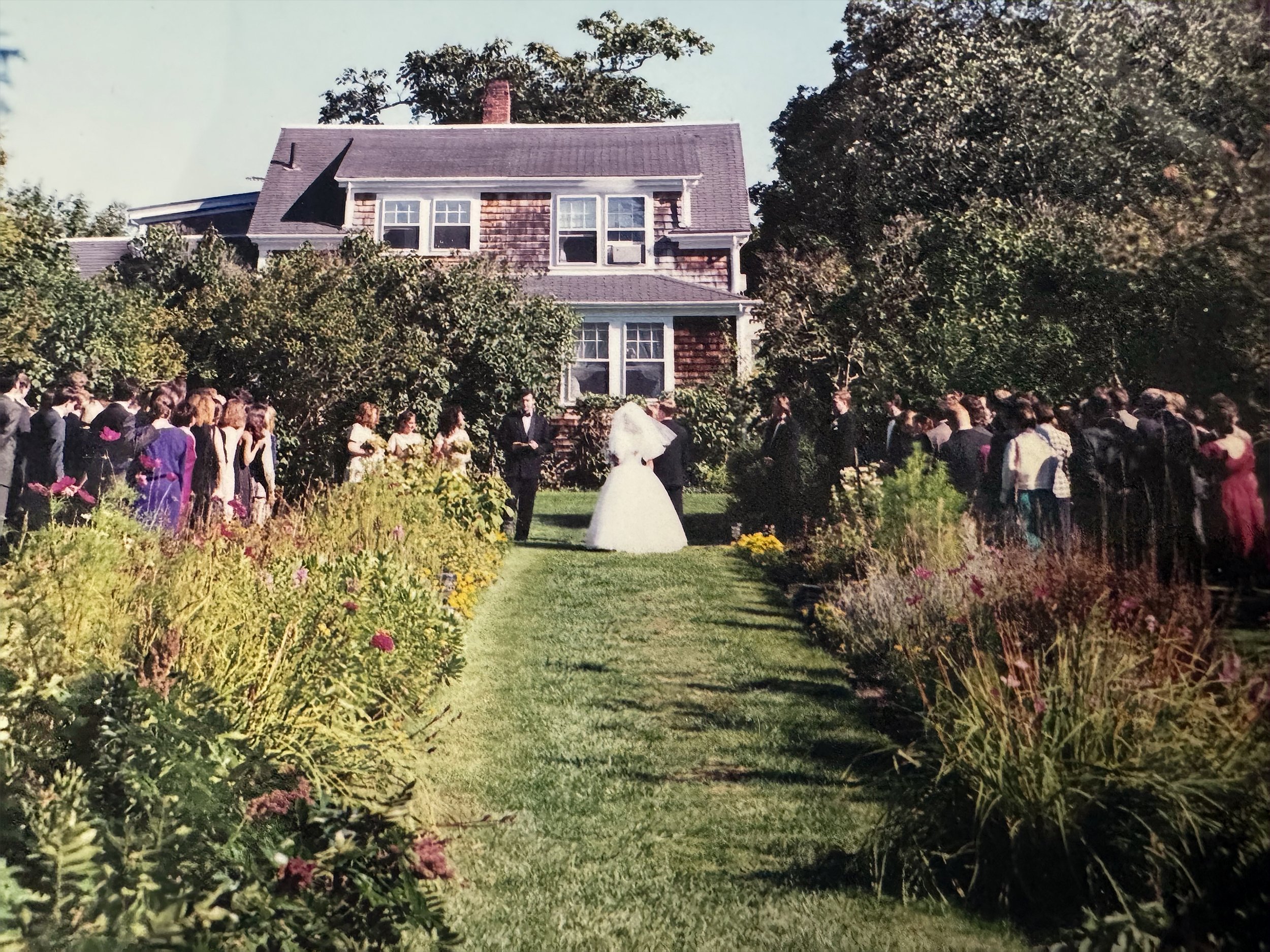 A wedding ceremony taking place outdoors in front of a large house with a gabled roof. The bride and groom are standing together under a small umbrella with a crowd of guests surrounding them, all dressed formally. The garden is lush with colorful fl