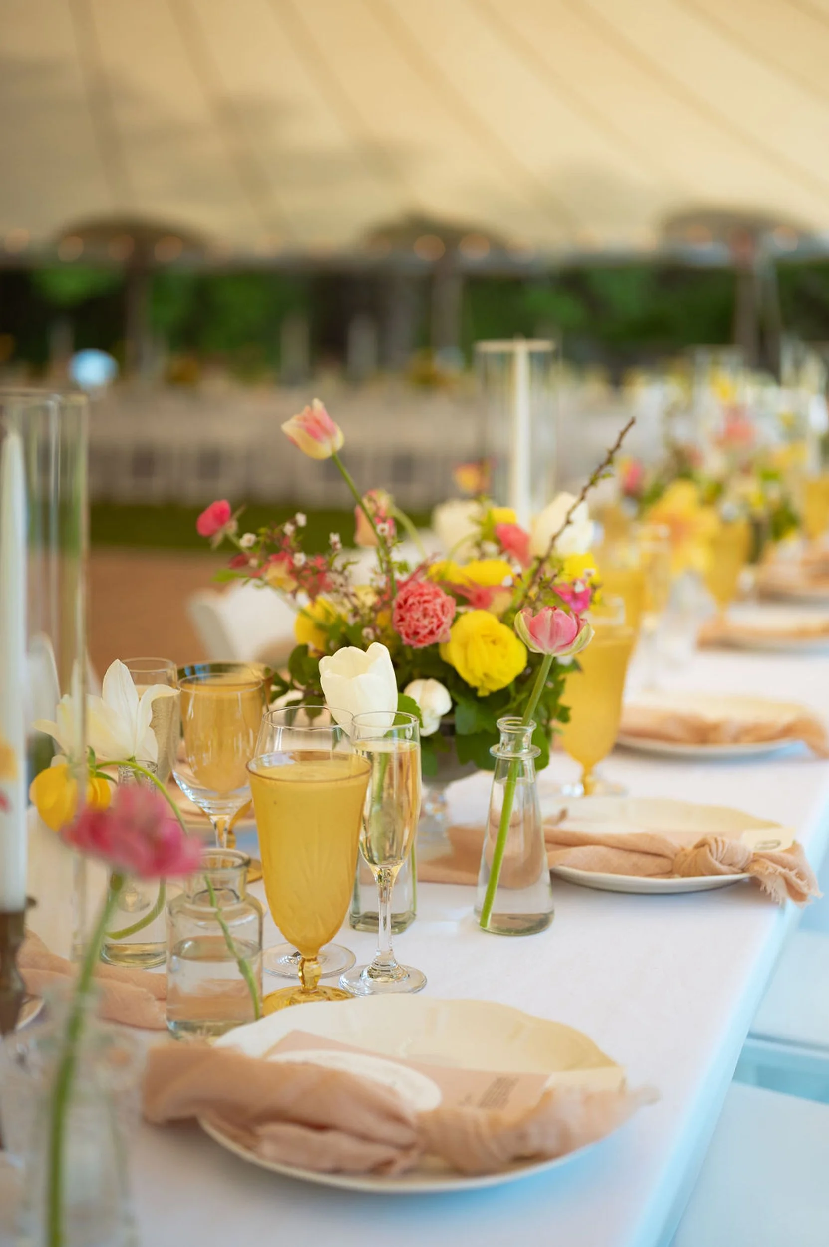 A table set for a wedding at Lambert's Cove Inn on Martha's Vineyard with colorful flower arrangements, glasses of yellow beverages, plates with napkins, and a blurry outdoor background.