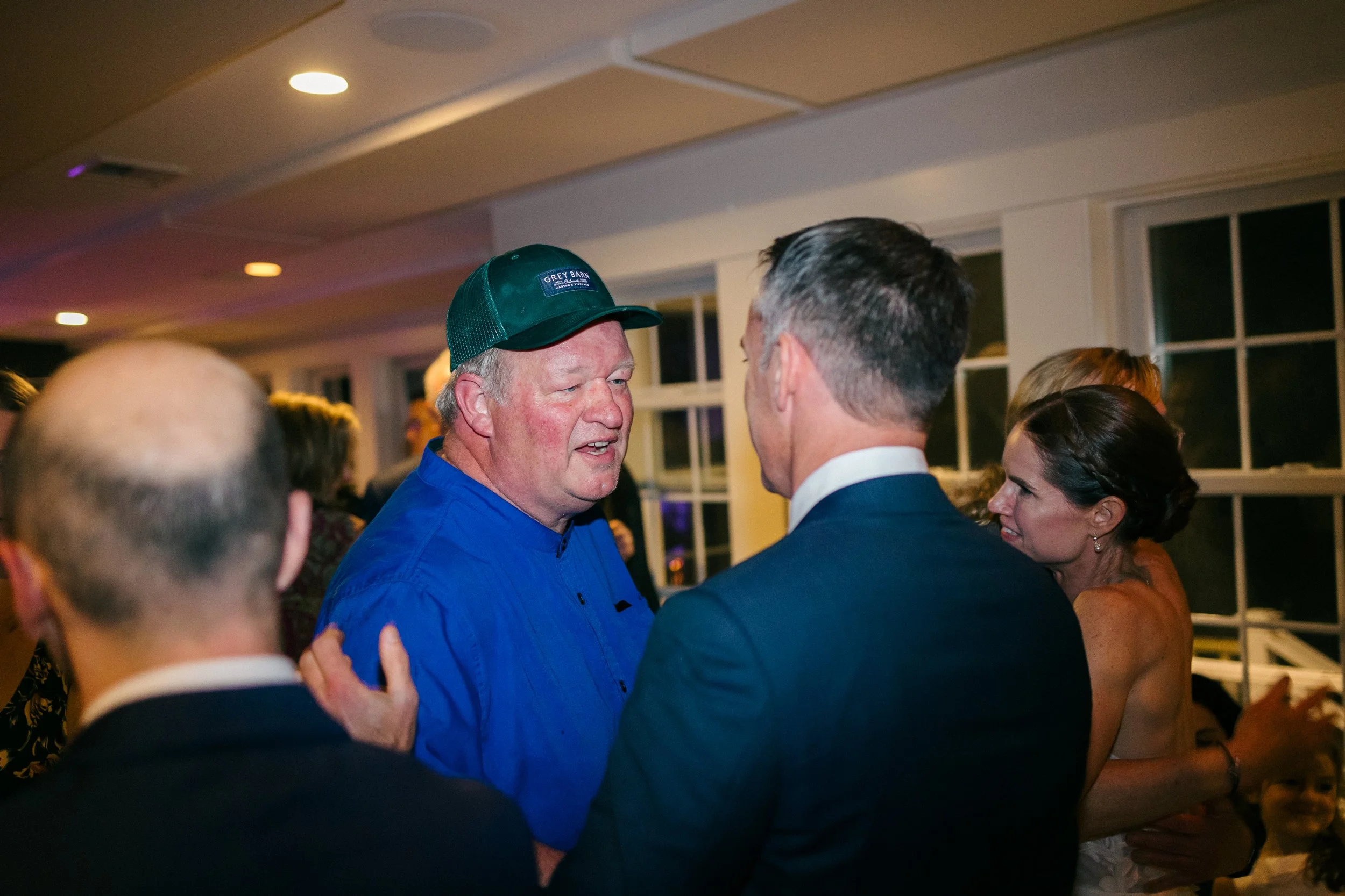 Two men are engaged in conversation at a social event. One man is wearing a green baseball cap and a blue shirt, while the other is dressed in a dark suit. Other people are visible in the background in a room with large windows and warm lighting.