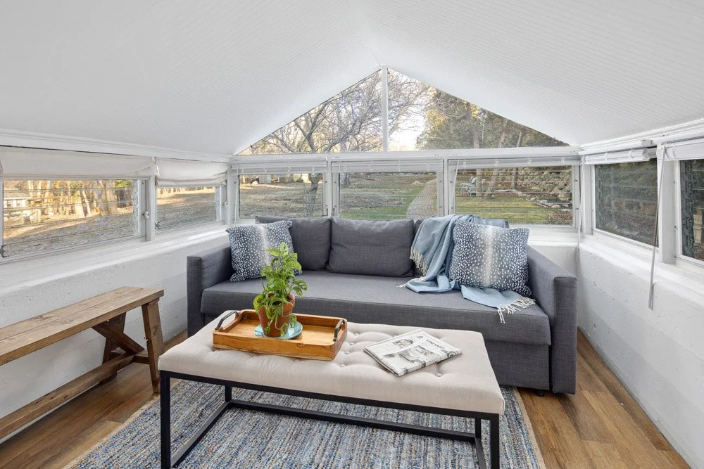 Enclosed porch with a gray couch, pillows, a blanket, a wooden bench, a cushioned ottoman with a tray and potted plant, a rug, and windows showing trees outside.