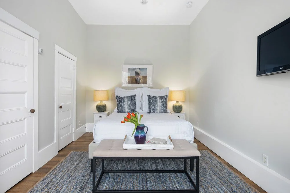 A bedroom with a bed, nightstands with lamps, a bench with flowers, and TV on the wall.
