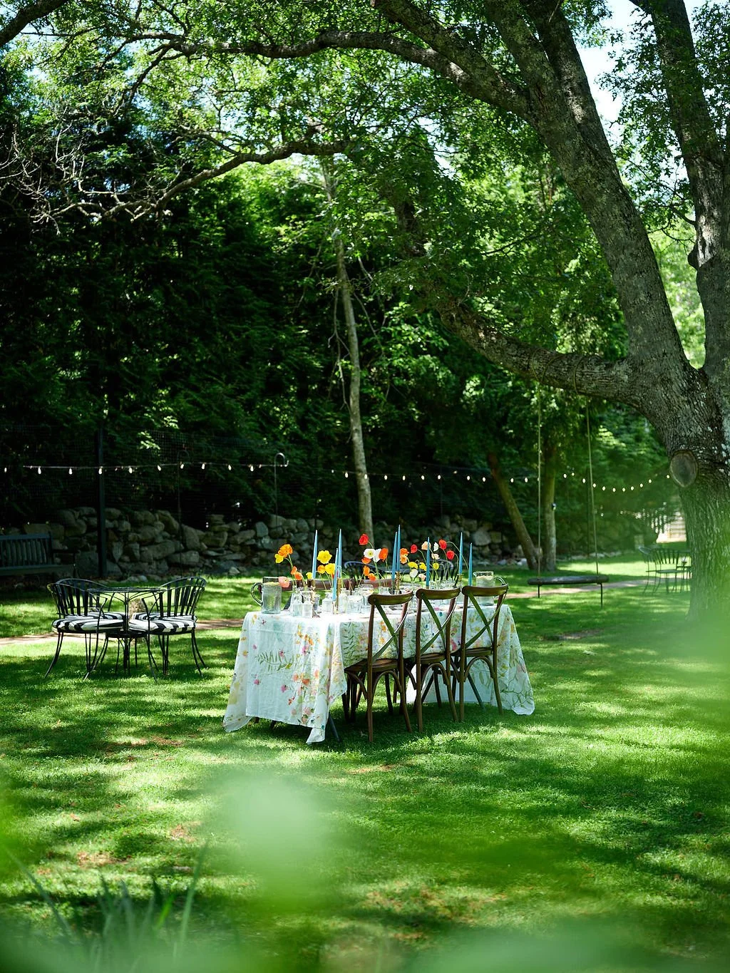 Outdoor garden scene with a table set for a meal, decorated with a floral tablecloth, colorful flowers, and tall blue candles. Surrounding the table are wooden and metal chairs, with trees and string lights overhead, creating a festive atmosphere.