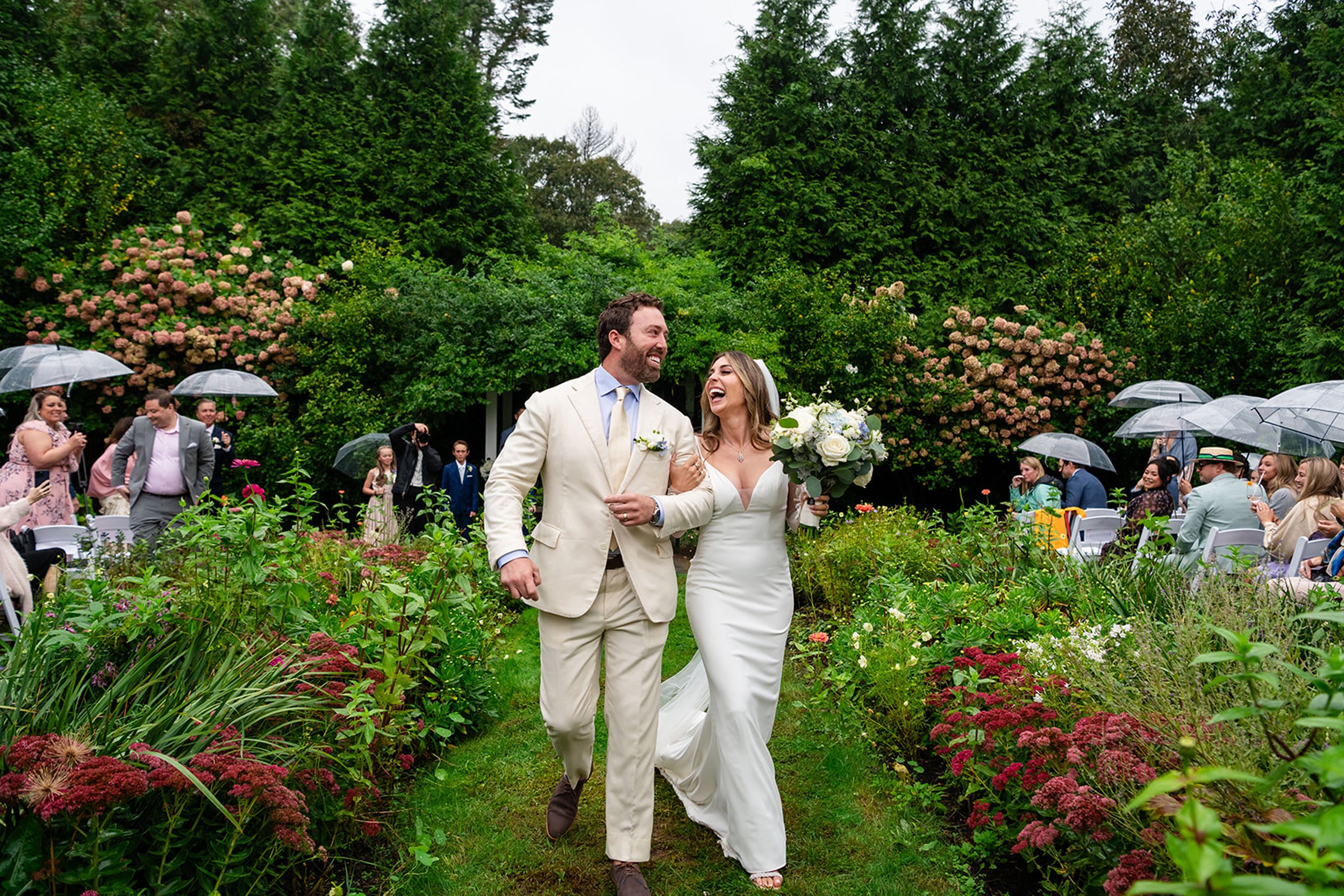 A newlywed couple walks arm-in-arm through a lush garden during their outdoor wedding reception, surrounded by friends and family holding umbrellas due to overcast weather.