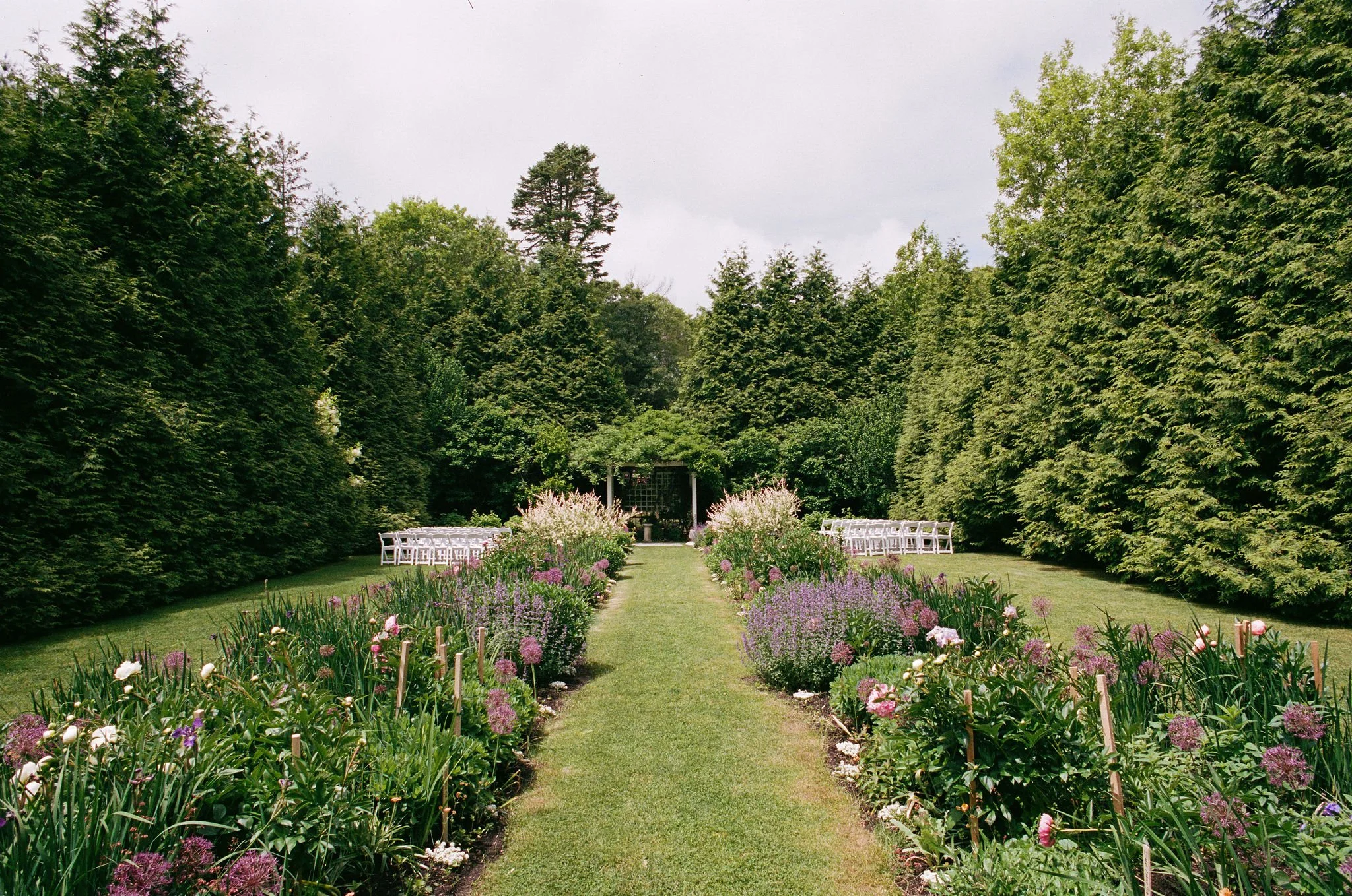 A beautiful outdoor garden at Lambert's Cove Inn on Martha's Vineyard, bordered by colorful flowers, tall green trees, and white chairs arranged along the sides for an event, with a small pavilion or gazebo in the background, under a cloudy sky.