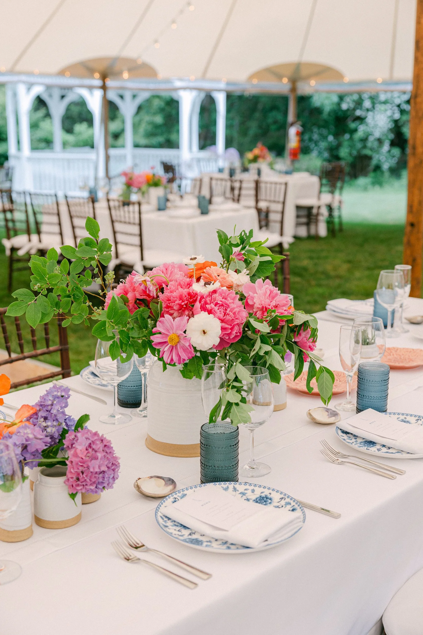 Elegant outdoor wedding reception setup with a decorated table featuring pink, purple, and white floral arrangements. The table is set with blue and white china plates, glasses, and silverware, under a white canopy with string lights. There are addit
