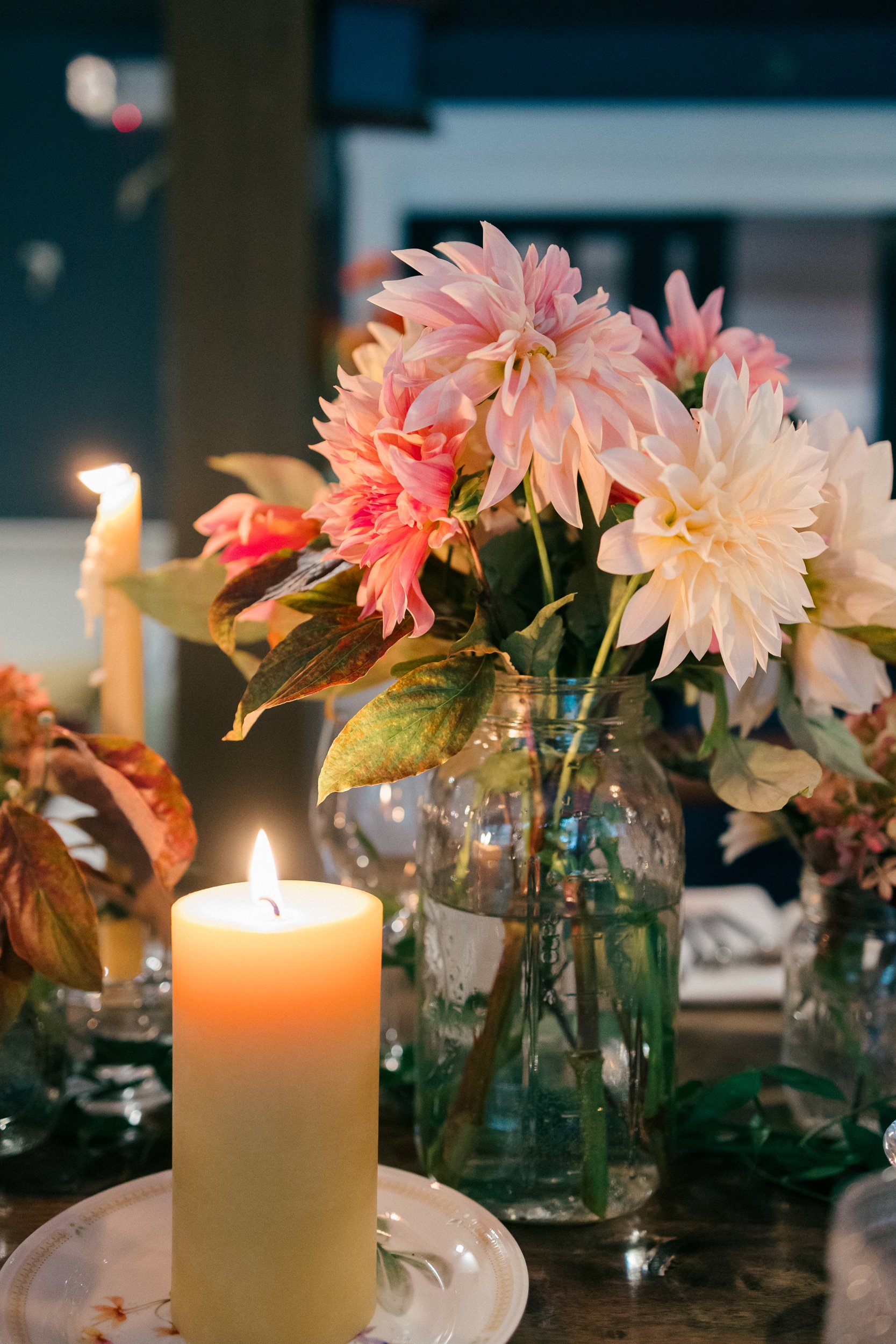 A table with pink and white dahlias in a glass vase, around a lit white candle and other decorations, during a wedding reception at Lambert's Cove Inn on Martha's Vineyard.